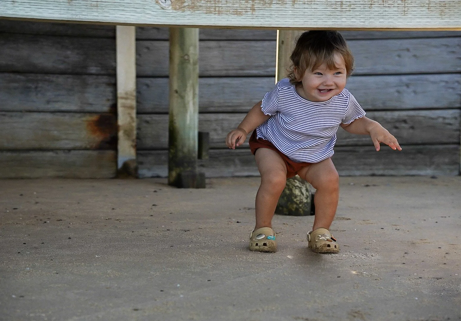 A young child with curly hair playing on a sandy beach, smiling and squatting near wooden posts and a wooden fence.