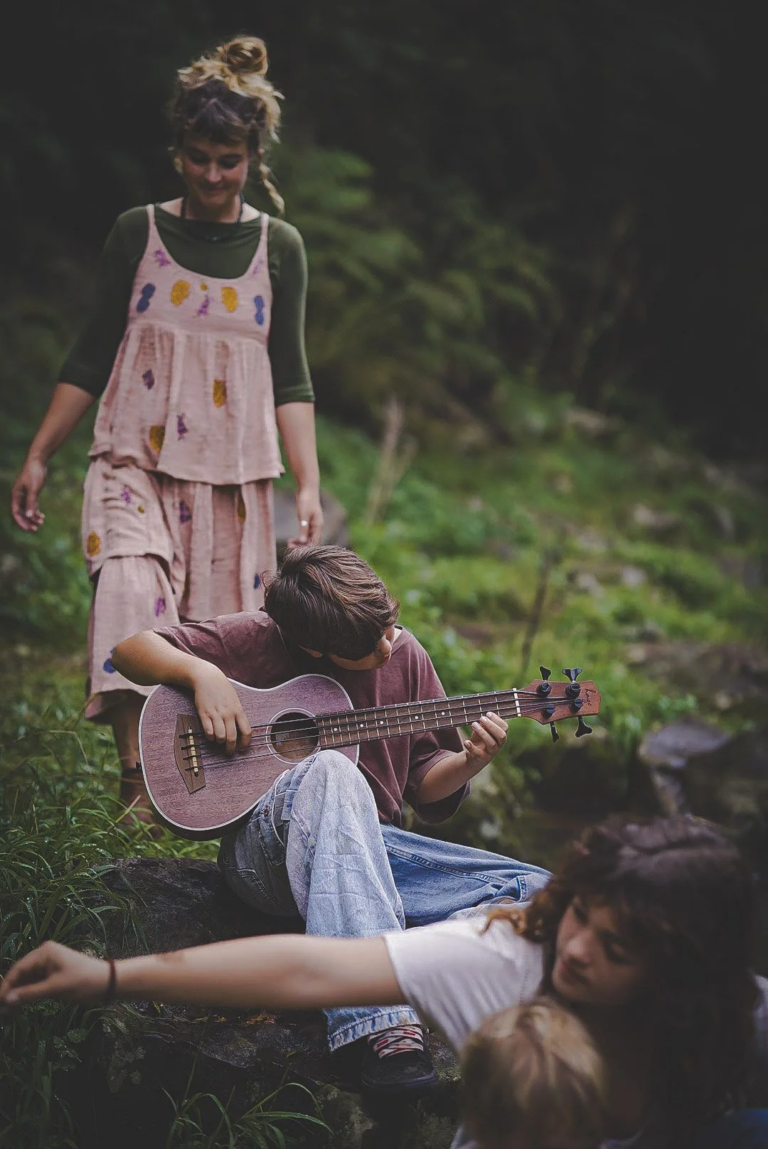 A young woman in a pink dress standing in a forest, watching a boy playing a ukulele while sitting on a rock. A woman is lying on the ground nearby, and a child with blonde hair is partially visible in the foreground.