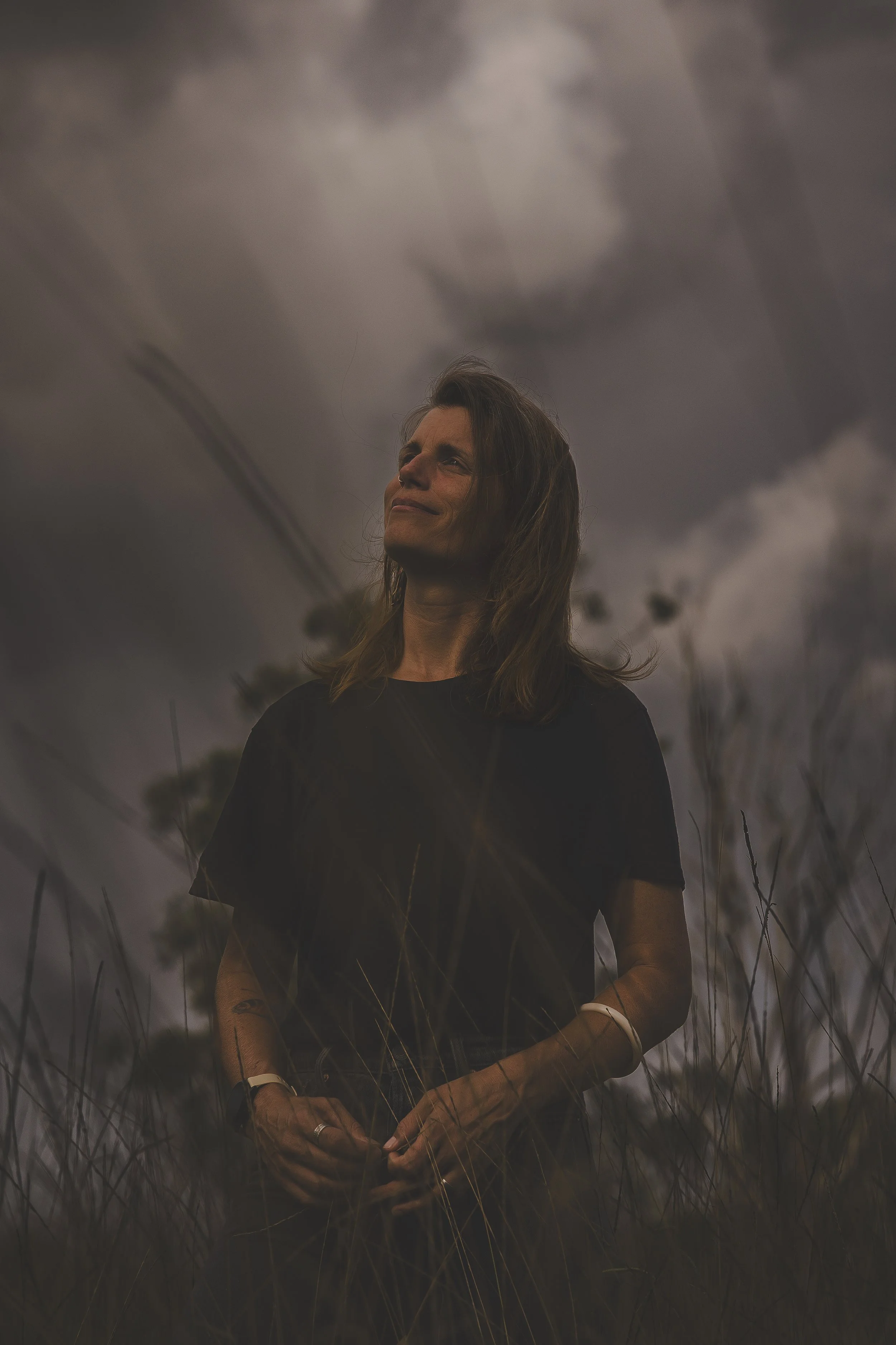 A woman standing outdoors in a field of tall grass under a cloudy, dark sky, looking contemplative with a slight smile.