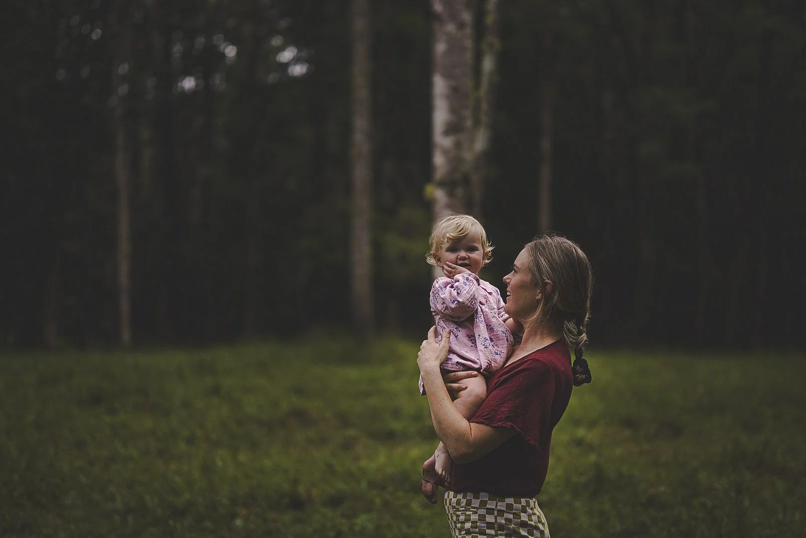 A woman holding a young girl outdoors in a forested area with tall trees in the background.