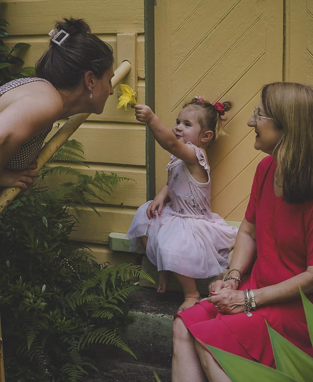 A young girl in a pink dress sitting on a porch, holding a yellow flower to the nose of an adult woman leaning towards her, while another woman in a red dress sits nearby, smiling.