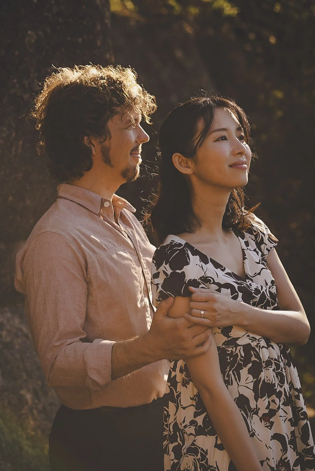 A man and woman standing outdoors during sunset, holding hands and looking contently into the distance. Engagement and LOVE.