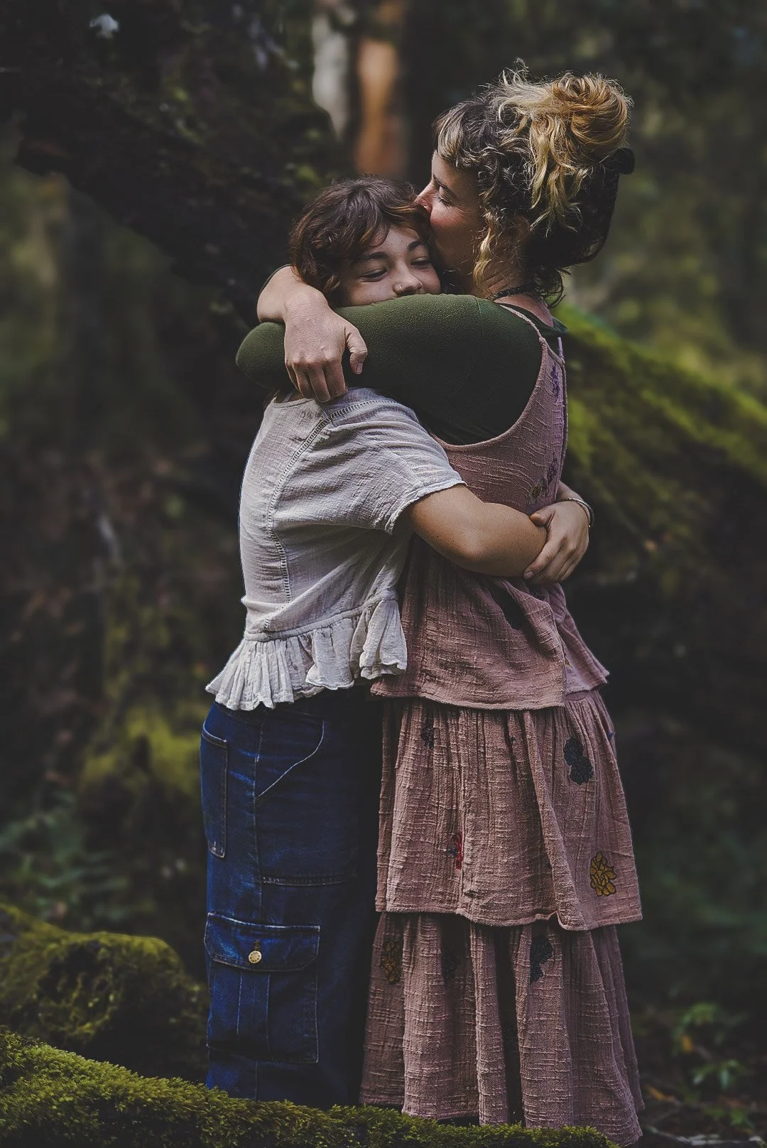 Mother and her daughter embracing in a forest, one with curly blonde hair giving a kiss on the forehead to a woman with short dark hair, both smiling.