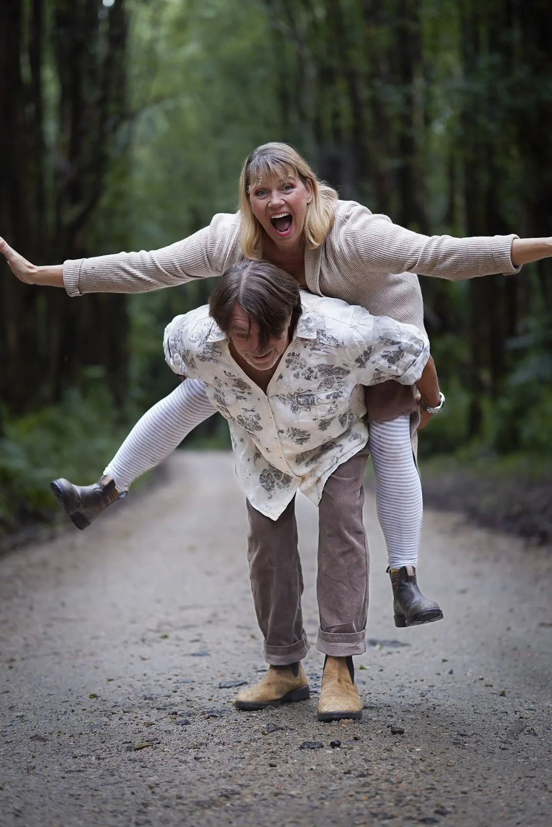 A woman is riding on a man’s back, spread arms in the air, smiling widely, as they walk down a forest trail. Husband and wife.