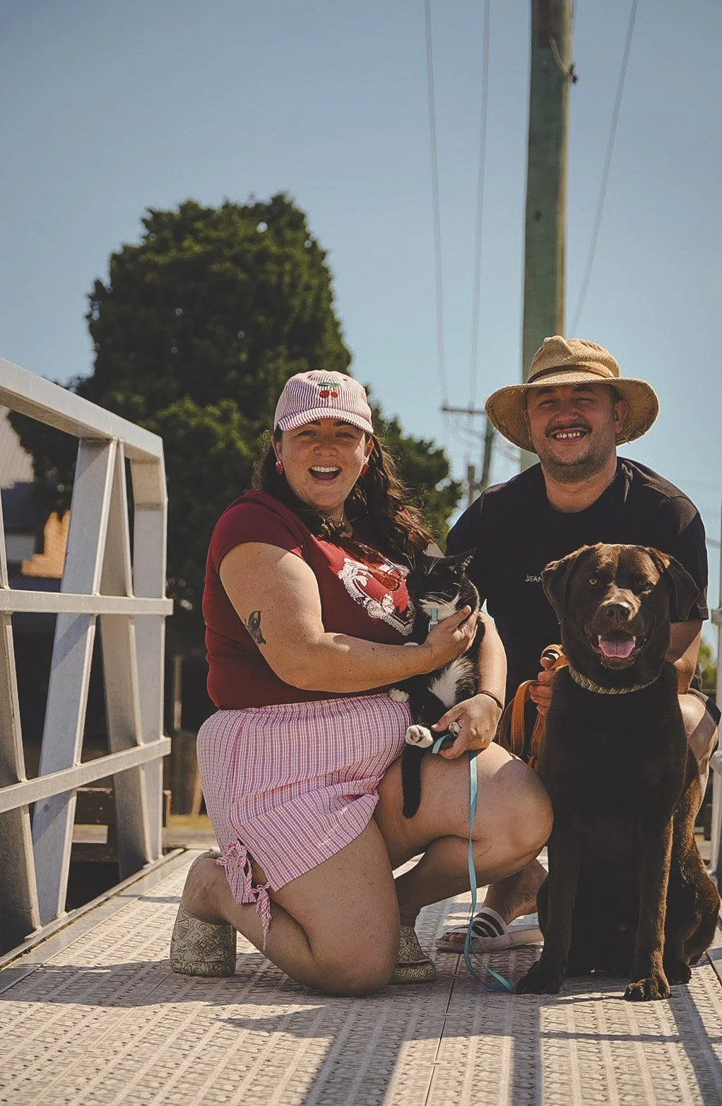 A smiling woman in pink striped shorts, red shirt, and a pink cap holds a black-and-white cat. Beside her, a man with a straw hat and a beard pets a large black dog. They are outdoors on a sunny day.