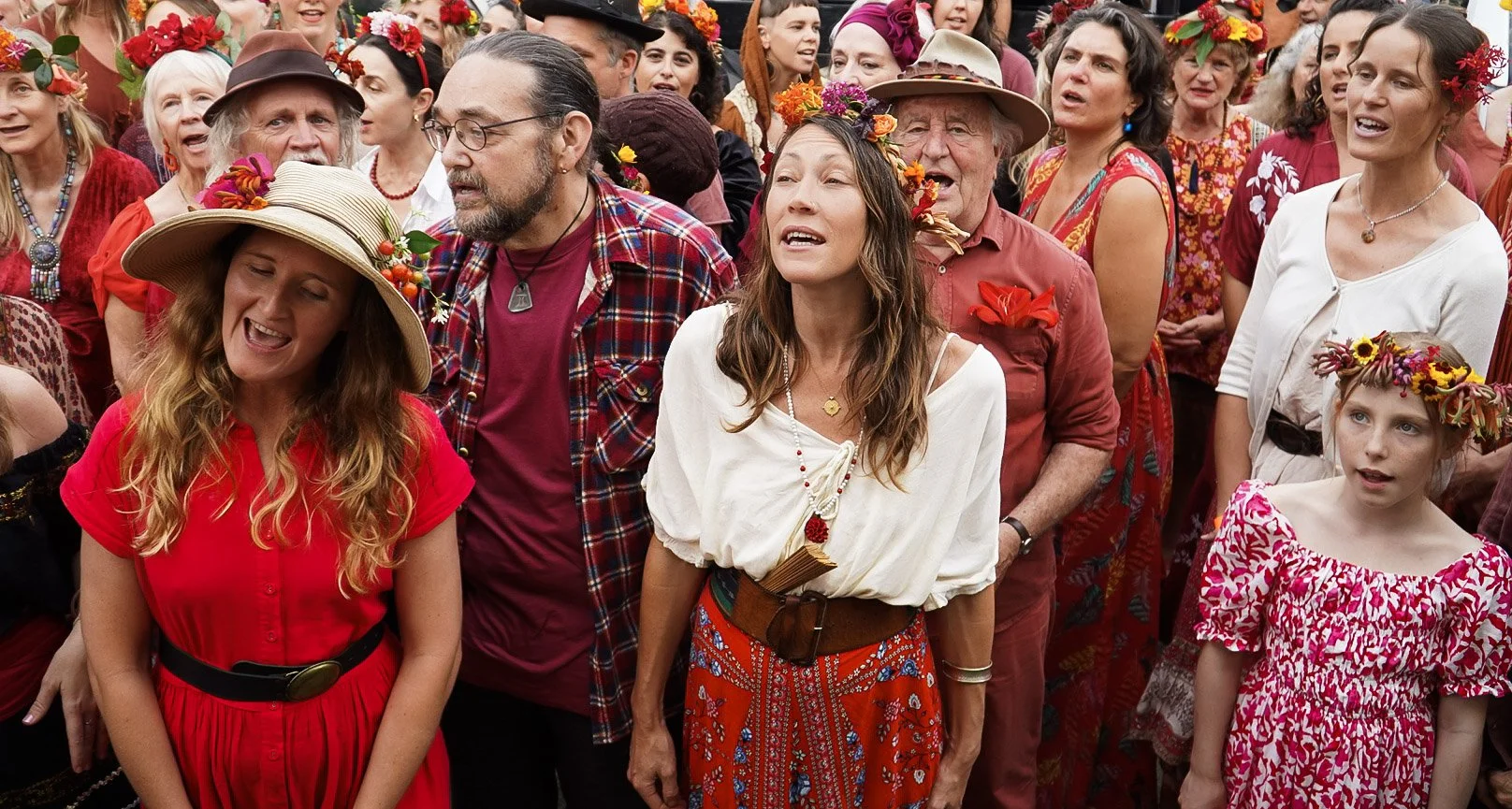 A crowd of people dressed in colorful, festive clothing, many wearing flower crowns and accessories, gathered outdoors for a celebration or festival.