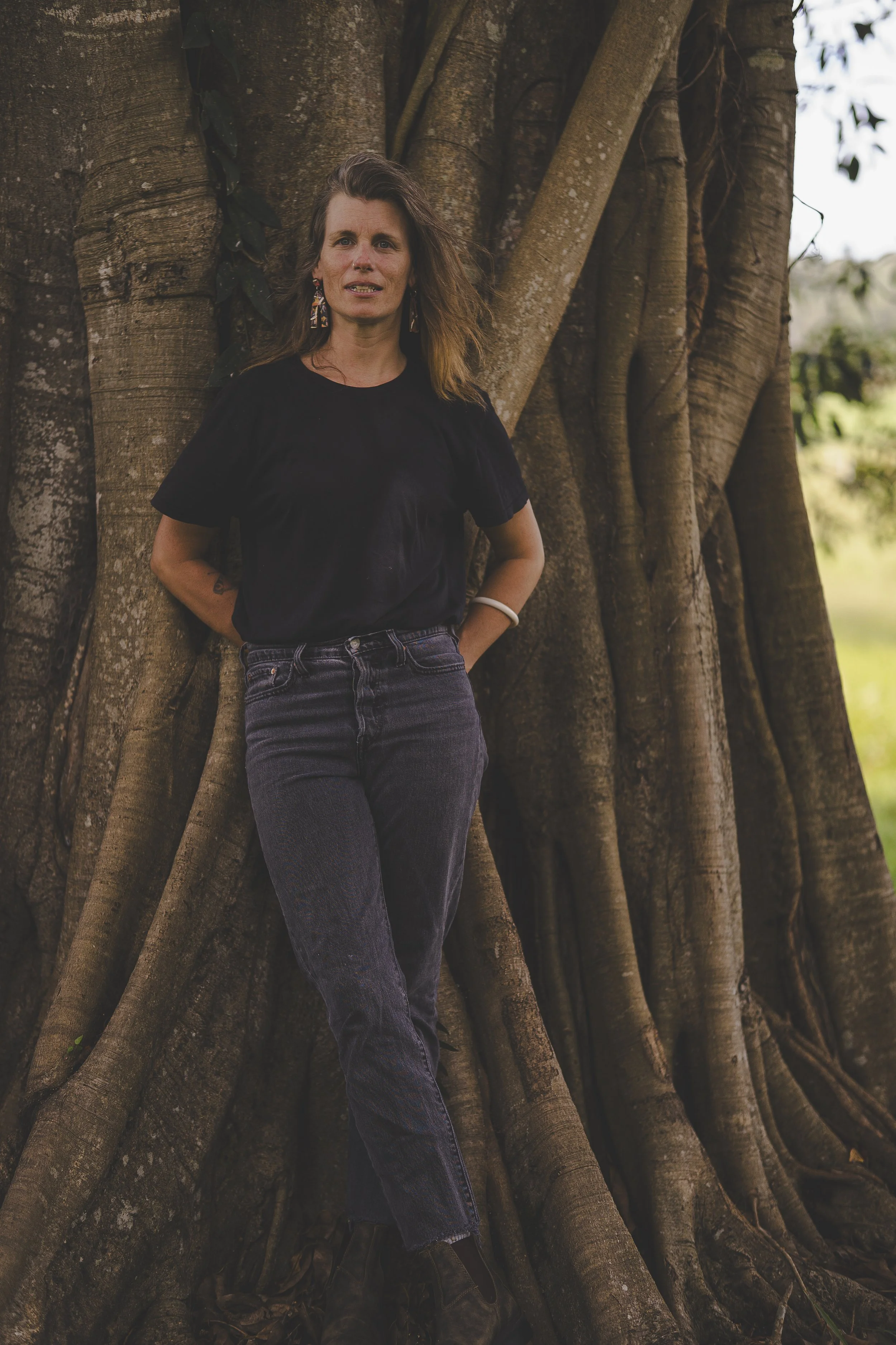 A woman with shoulder-length hair leaning against a large tree trunk outdoors, wearing a black T-shirt, dark jeans, and earrings.