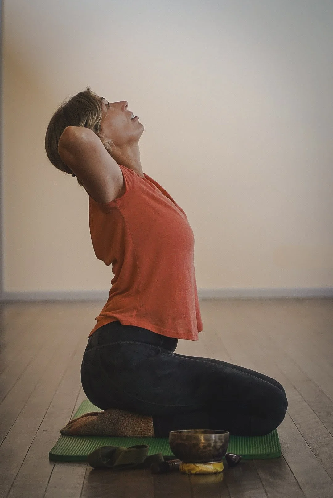 Woman practicing yoga on a green mat, kneeling with arms behind her head, in a room with a light-colored wall, with a singing bowl, scarf, and stones in front of her.