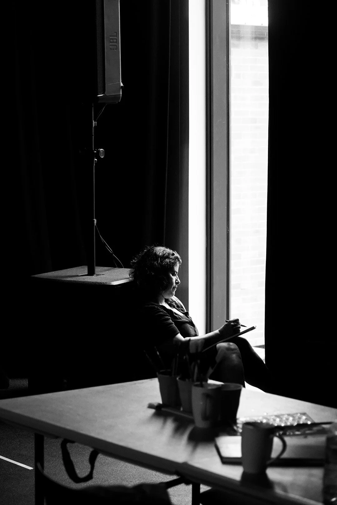A woman sitting by a window, writing or drawing on a notepad, inside a dark room with a large speaker and a table with cups and stationery.