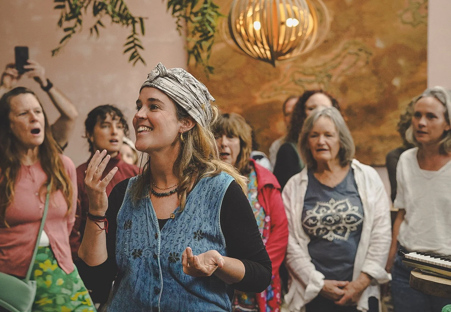 A group of women gathered indoors, with one woman in the foreground smiling and gesturing while others around her listen or sing. The background features warm-colored walls and decorative lighting.