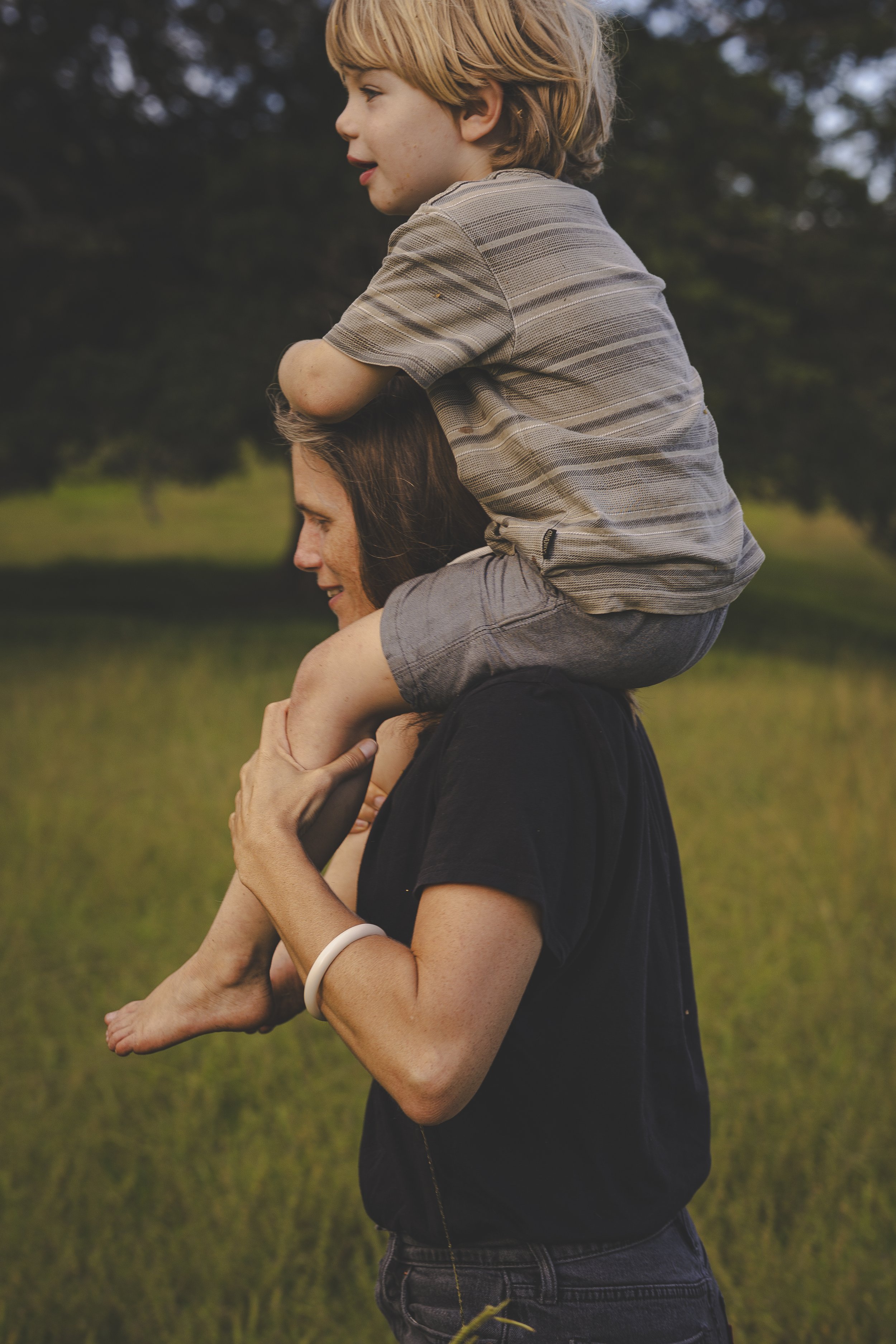 Woman carrying a young boy on her shoulders outside in a grassy field with trees in the background.