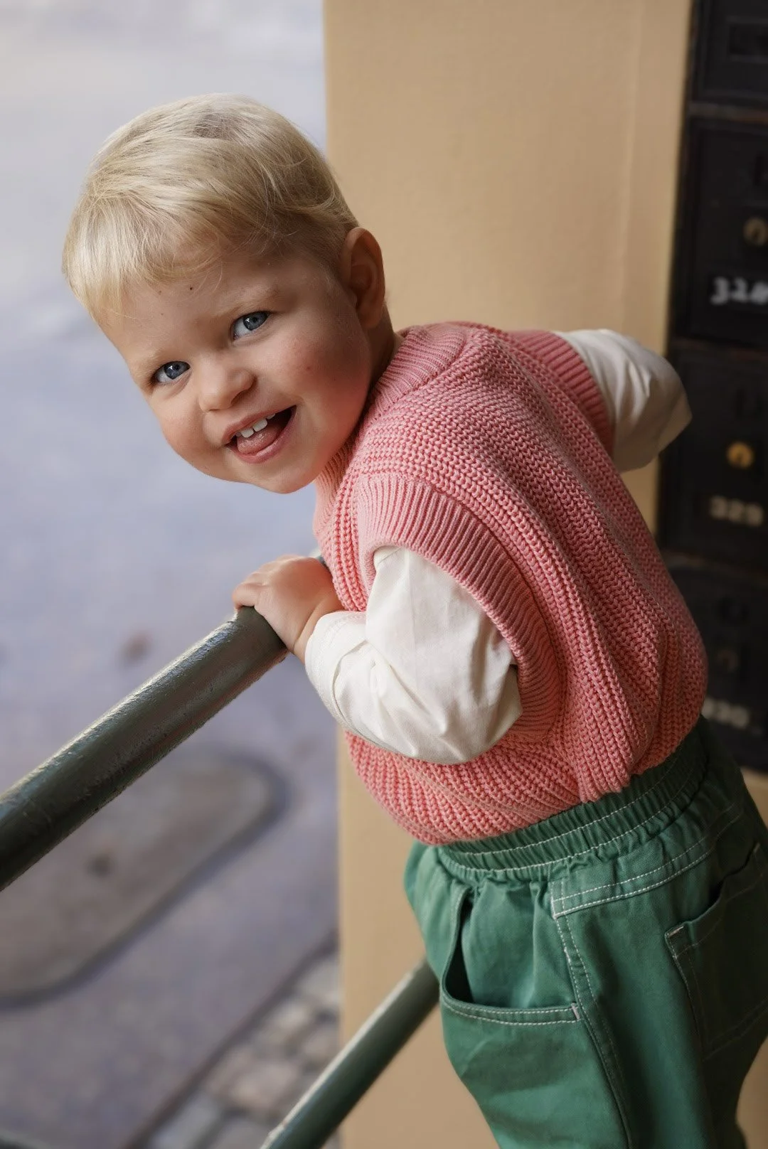 A young boy with blonde hair and blue eyes smiling while leaning on a railing indoors.