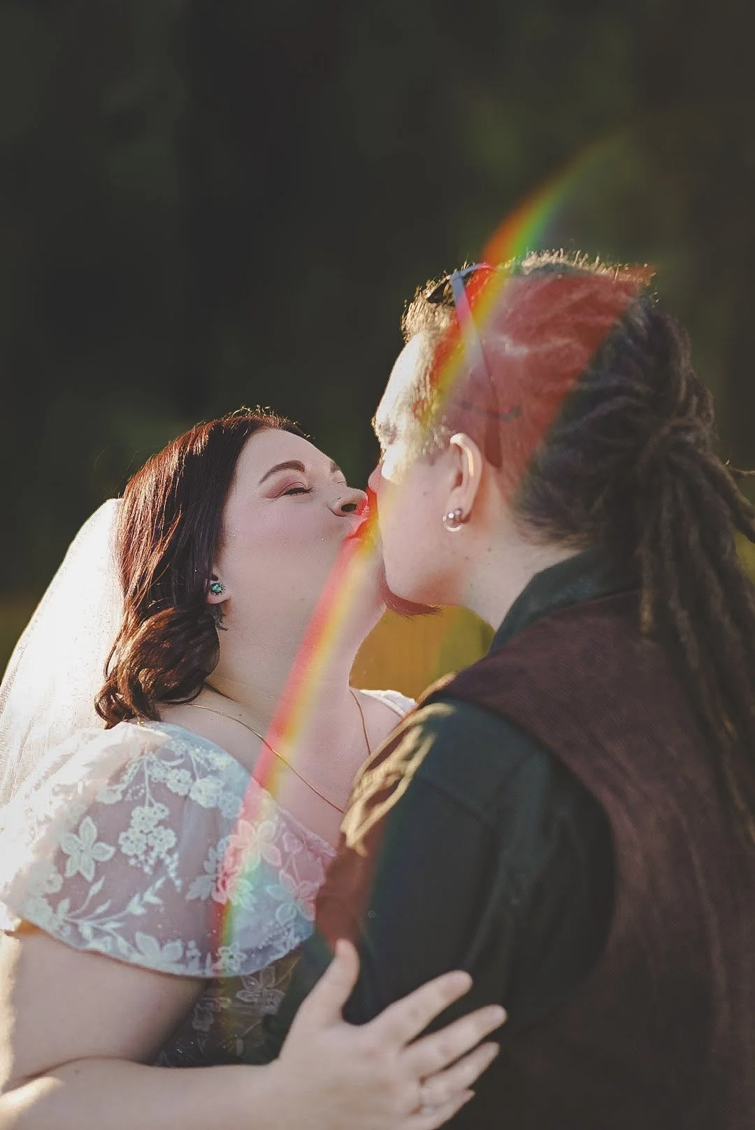 A woman in a wedding dress and a man are kissing outdoors, with a rainbow visible in the sunlight.