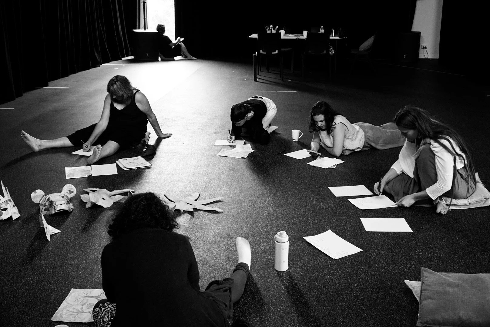 Five women sitting and lying on a dark carpeted floor, engaged in writing or reading papers, with some objects scattered around, in a dark room with a bright window in the background.