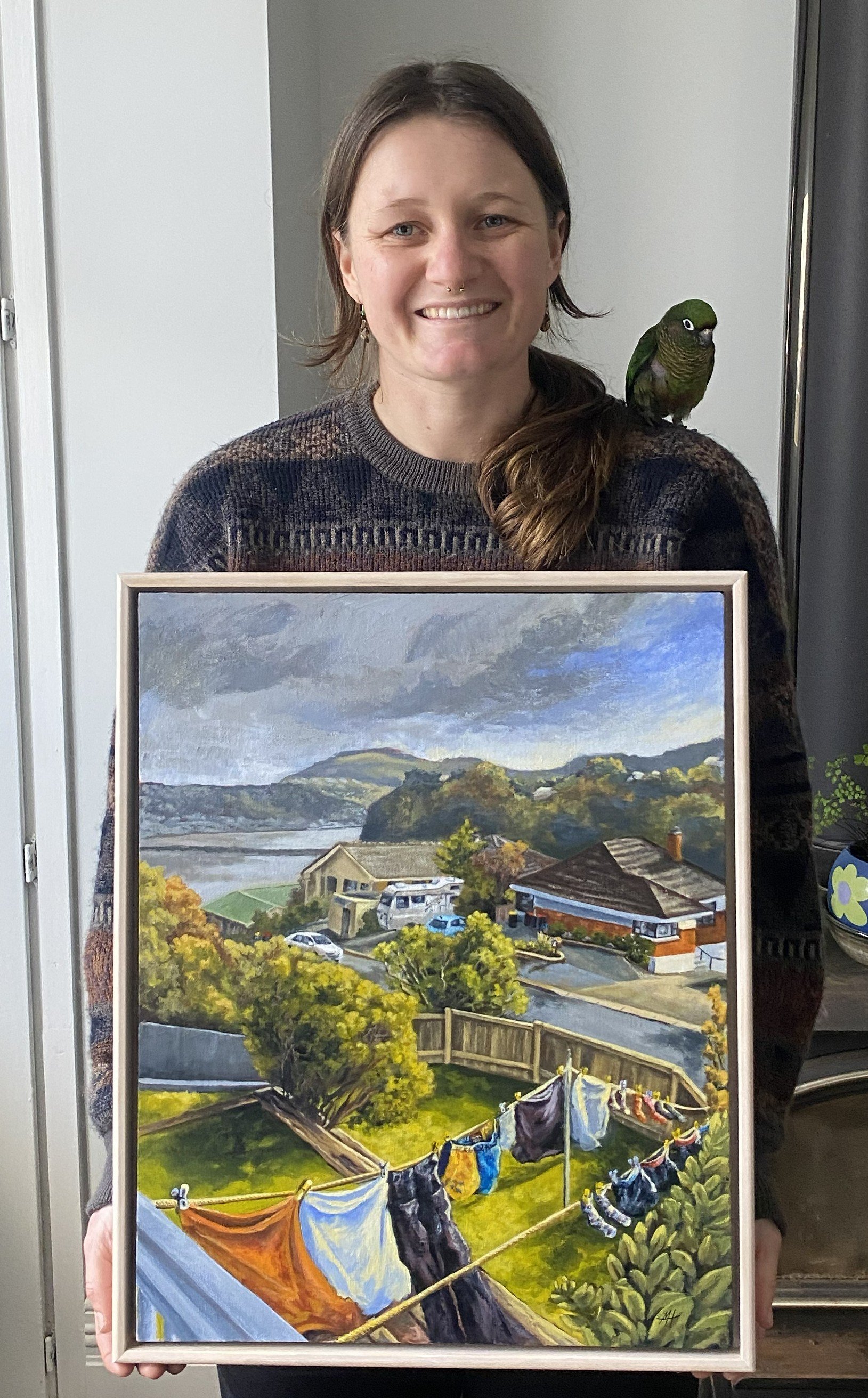 A woman with long brown hair and a septum piercing holding a framed painting of a neighborhood scene with houses, trees, and laundry drying on a clothesline in a backyard.