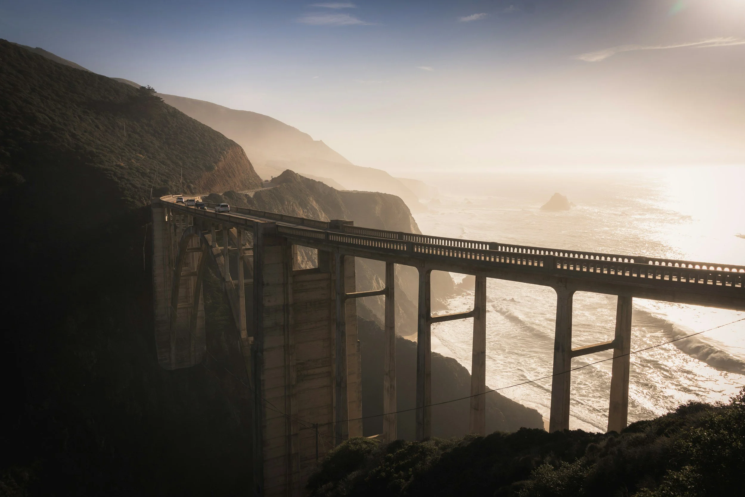 A scenic view of a coastal highway bridge spanning a deep canyon with waves crashing below, during sunset.