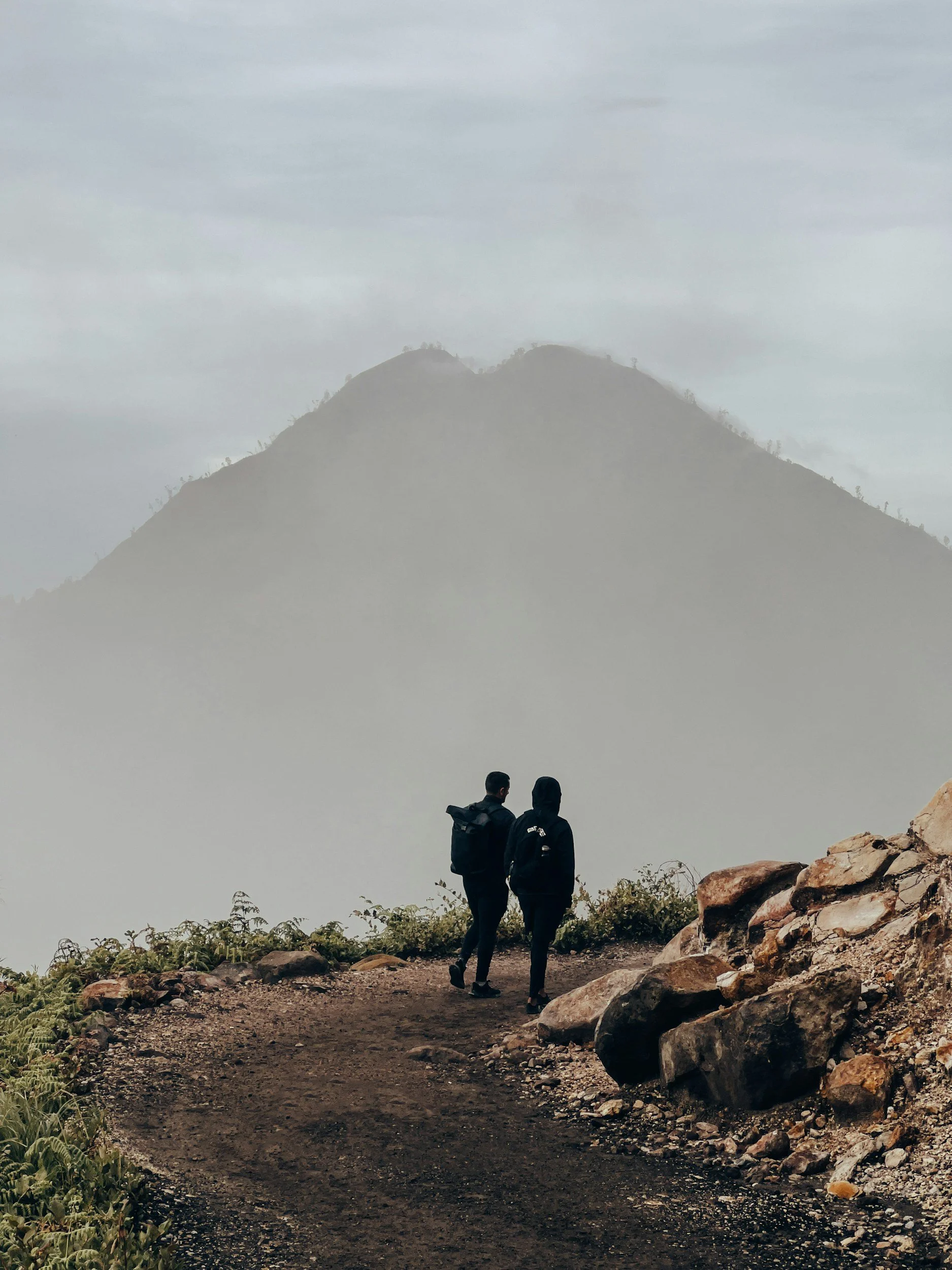 Two hikers walking along a dirt trail on a mountain with a foggy landscape and a large mountain in the background.