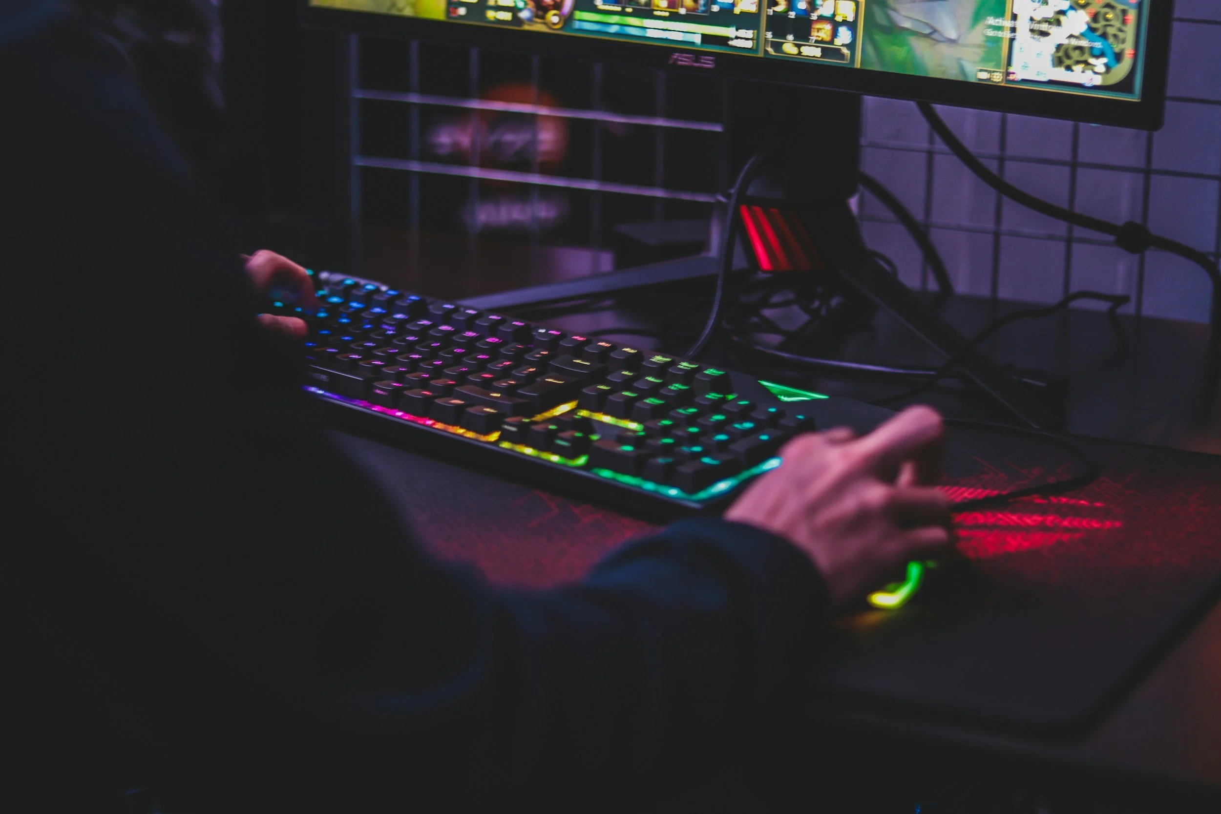 Pessoa jogando computador com teclado e mouse com iluminação RGB em ambiente de sala de jogos.