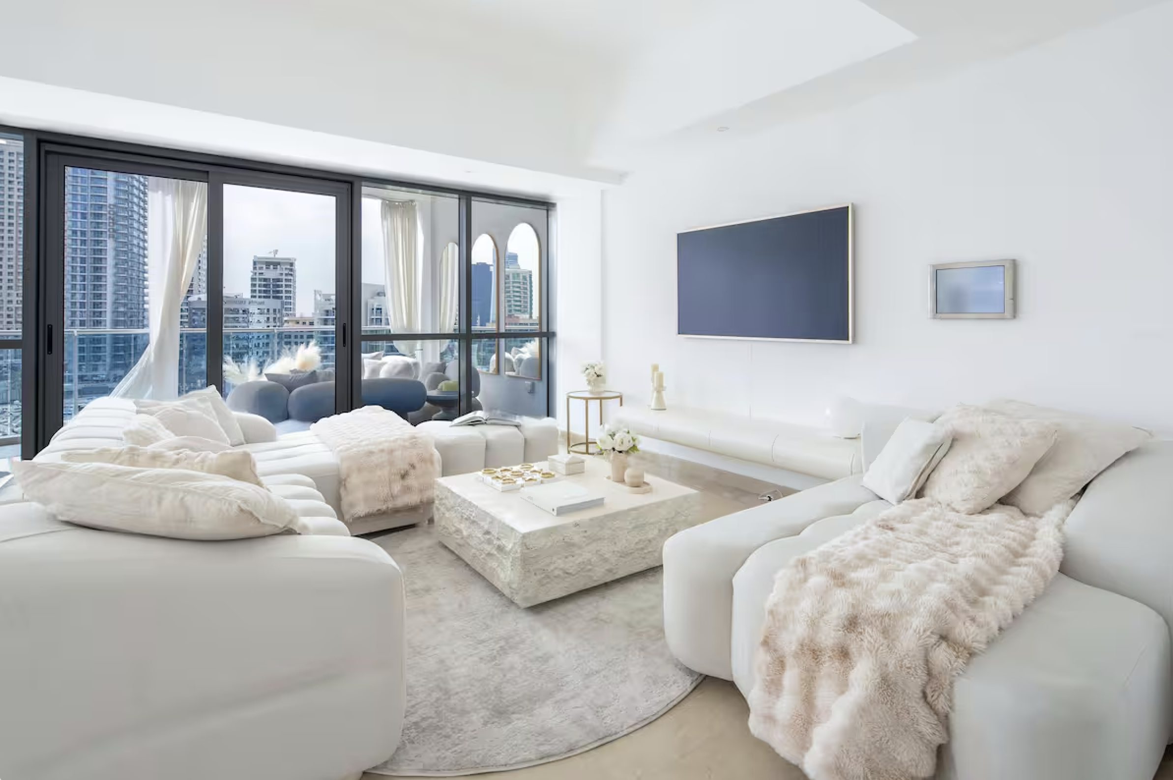 Bright modern living room with white sofas, plush blankets, a stone coffee table, and large windows with city views.