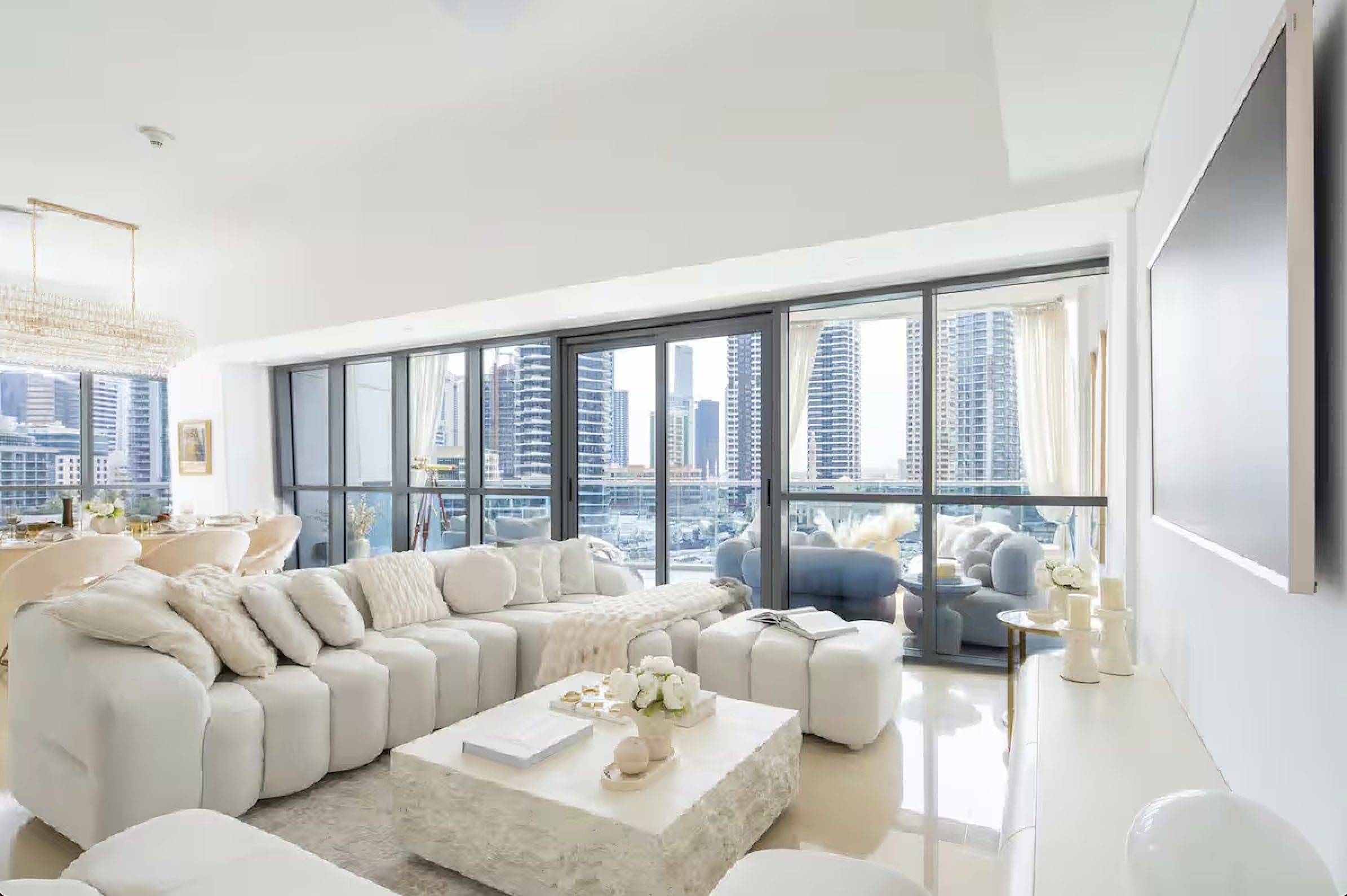 Modern living room with large white sectional sofa, glass sliding doors leading to balcony with cityscape view, and light-colored decor.