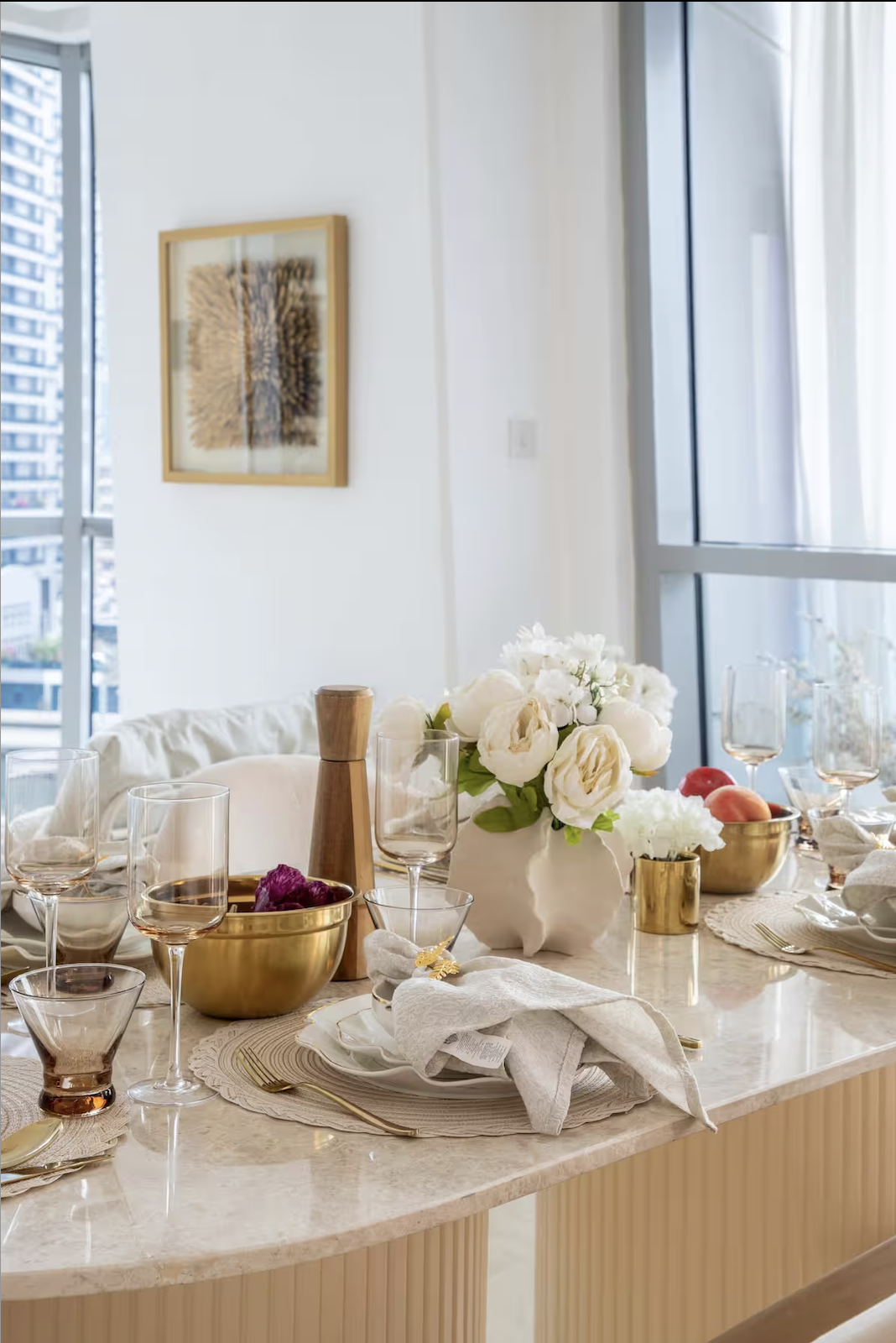 A dining table set for a meal with white flowers in a vase, gold and clear glasses, gold utensils, a wooden pepper shaker, bowls with fruits, and cloth napkins. The room has large windows with sheer curtains, and a framed artwork on the wall.