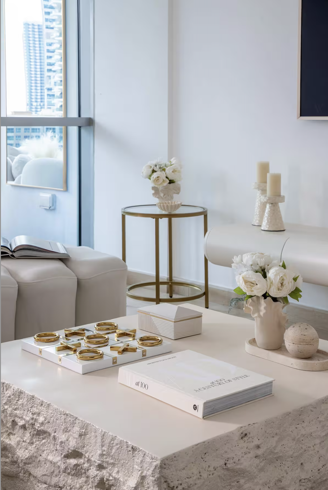 Modern living room with white and gold decor, a white table with gold rings and books, flowers in vases, candles, and a large window showing a cityscape.