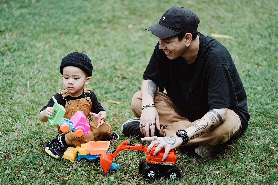 A young man with tattoos, black shirt, beige pants, and a black cap playing with a toddler girl on grass. They are surrounded by colorful toy construction vehicles, and the young man is smiling and engaging with the girl.