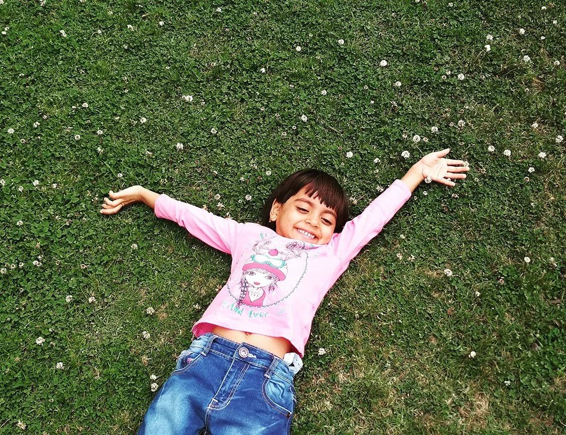 A young girl lying on her back on the grass with her arms outstretched, smiling.