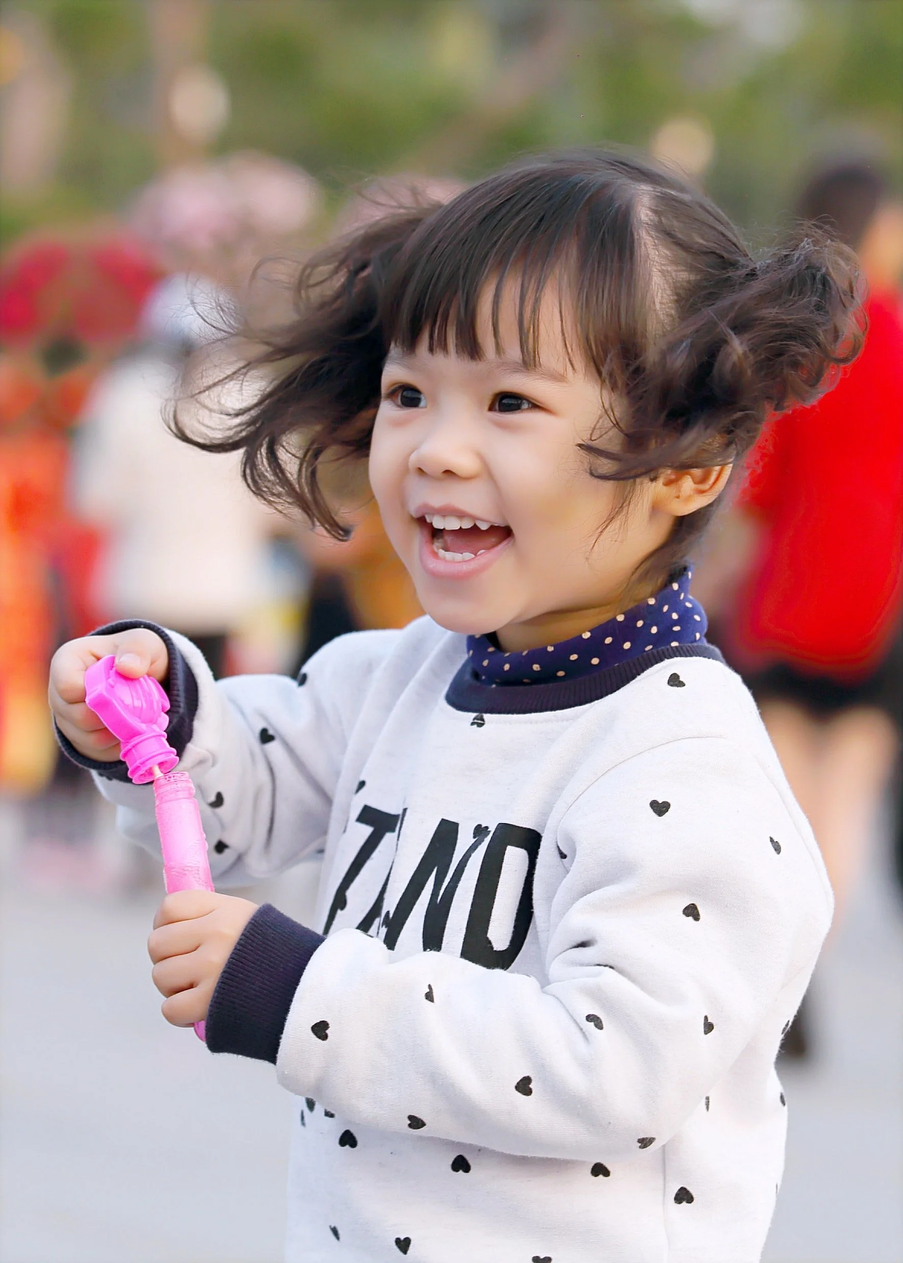 A young girl with curly hair, smiling and holding a pink bubble wand, outdoors at a festive event.