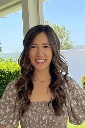 Young woman with long, wavy hair smiling outdoors on a sunny day.