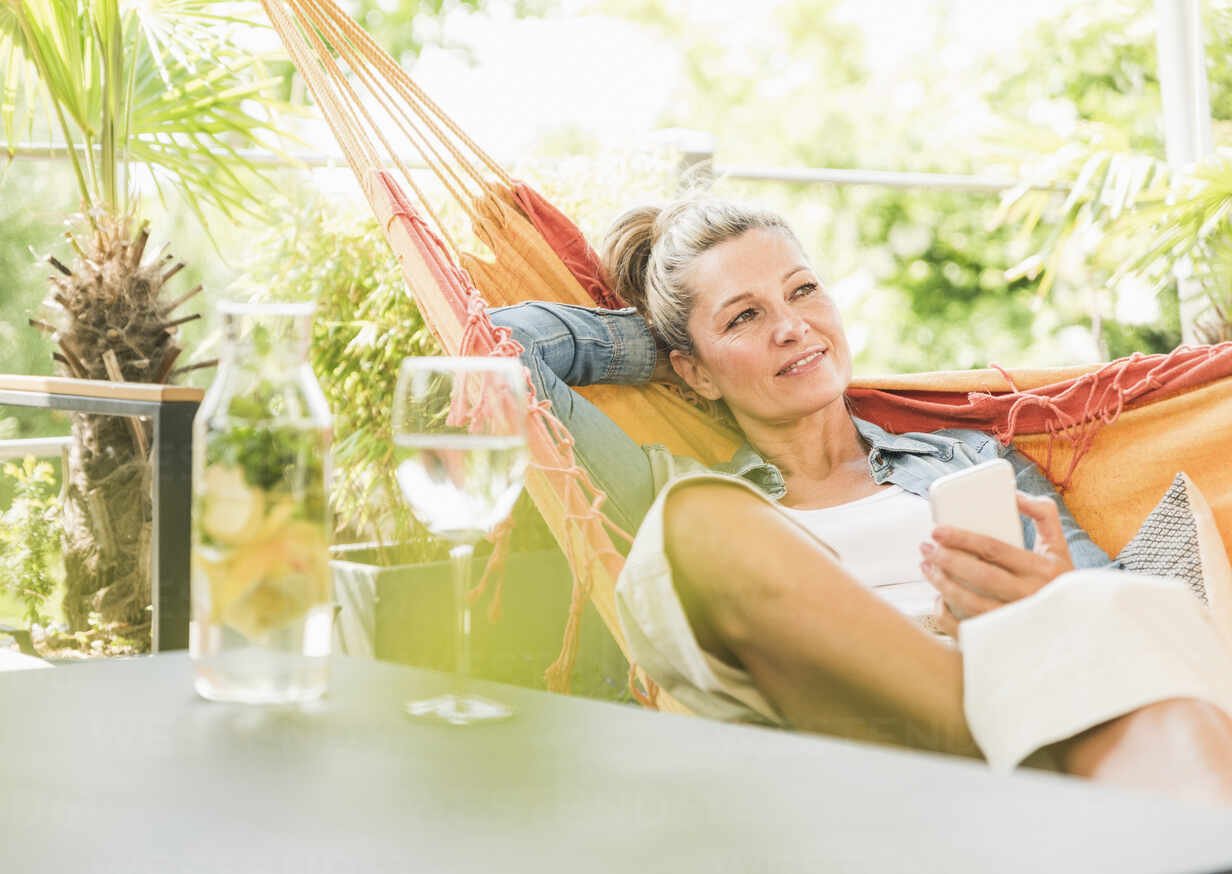 A woman relaxing on a hammock outdoors, holding a smartphone, with a glass of water and plants nearby.