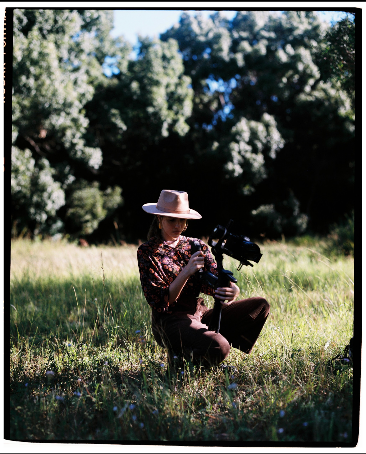A woman sitting on the grass in a park, holding a camera with a stabilizer, wearing a wide-brimmed hat and floral blouse, surrounded by trees and sunlight.