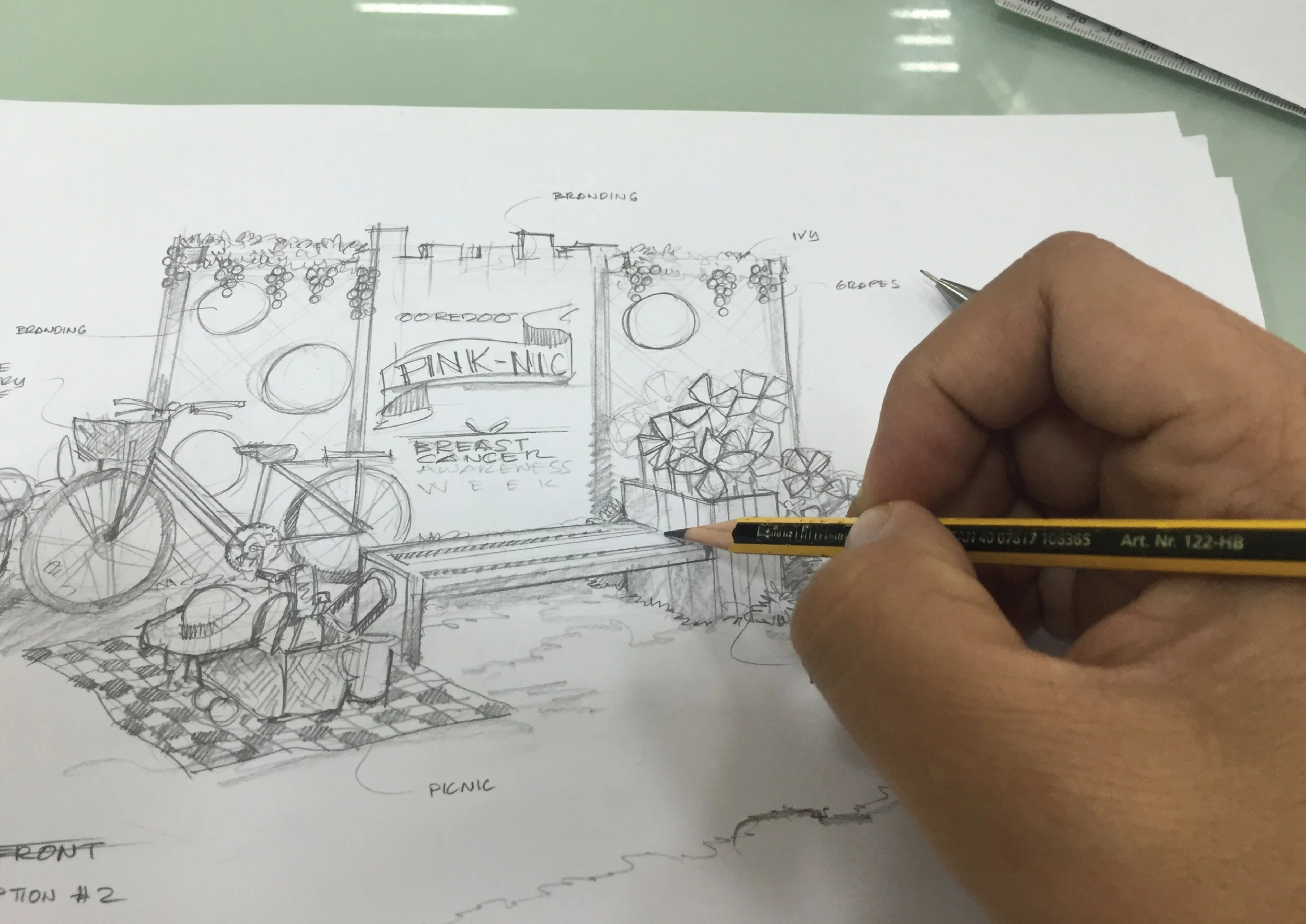 Hand sketching an outdoor garden scene on paper with a pencil. The scene includes a bicycle, picnic area, plants, and banners with text.