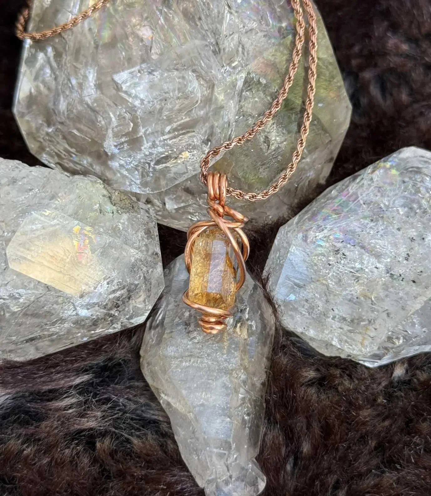 A necklace with a pendant made of a wire-wrapped yellow crystal, resting on large clear quartz crystals on a dark furry surface.