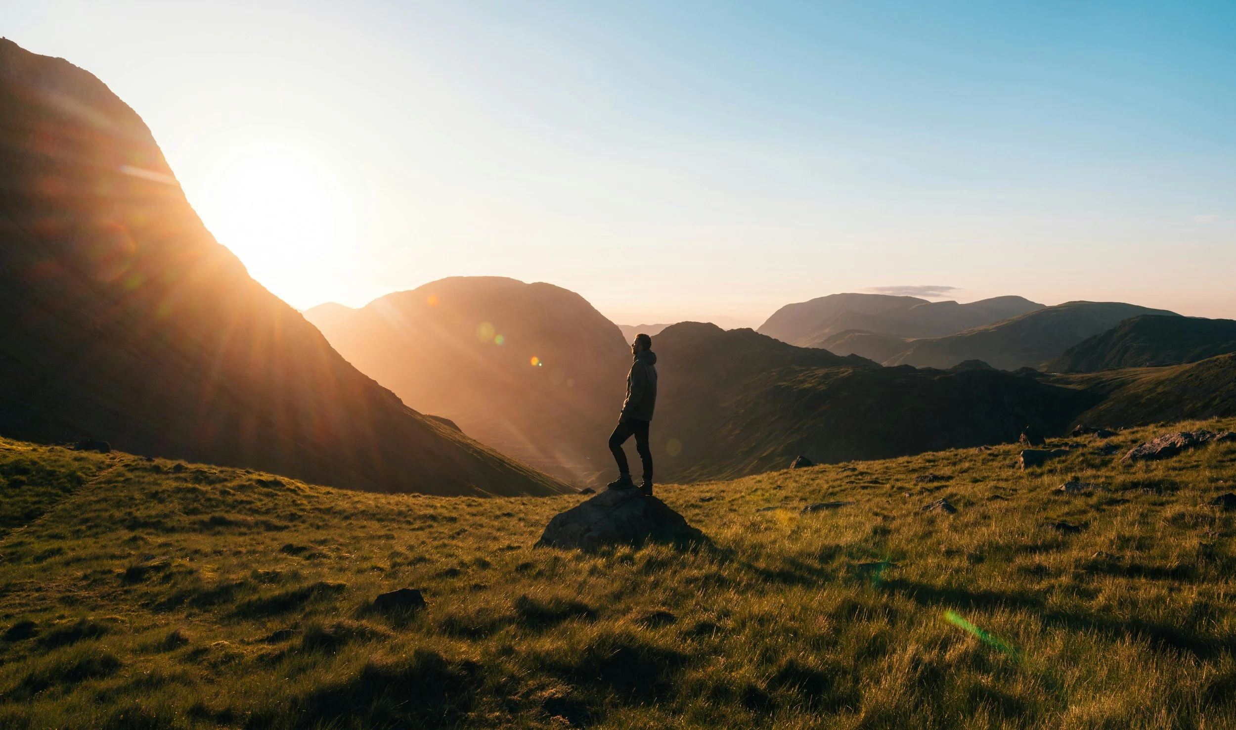 A person standing on a rock in a grassy valley during sunset, surrounded by mountains.