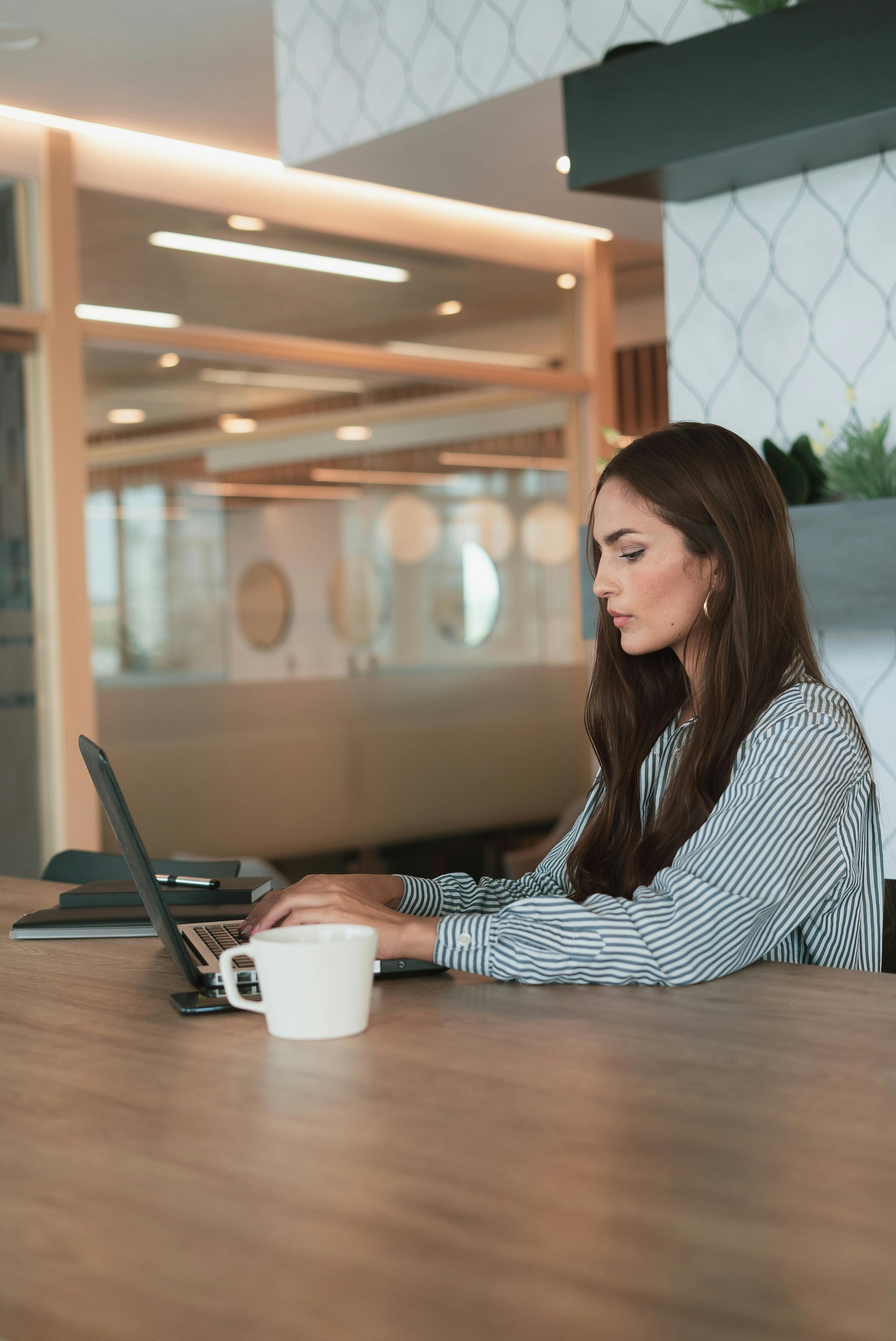 A young woman with long brown hair, wearing a striped shirt, working on a laptop at a wooden table in a modern office or cafe setting, with a white mug and notebooks nearby.