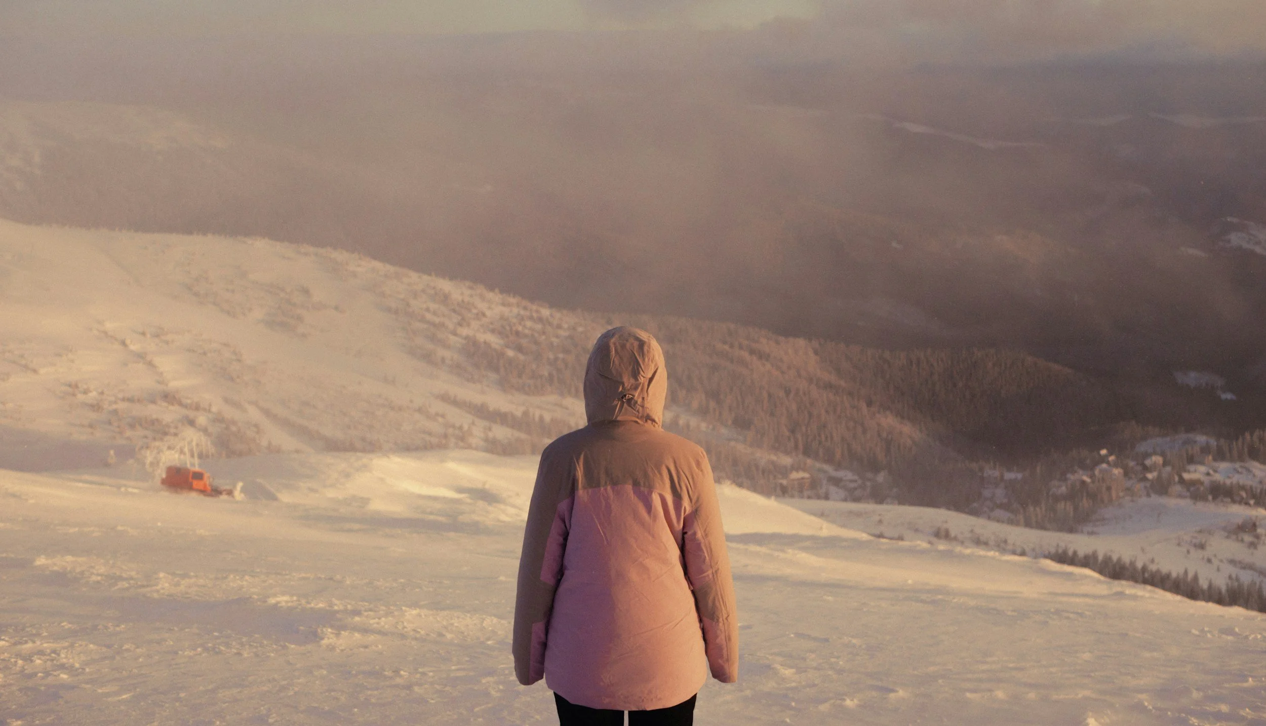 Person standing on snow-covered ground overlooking a mountainous landscape with snow and trees, during sunset