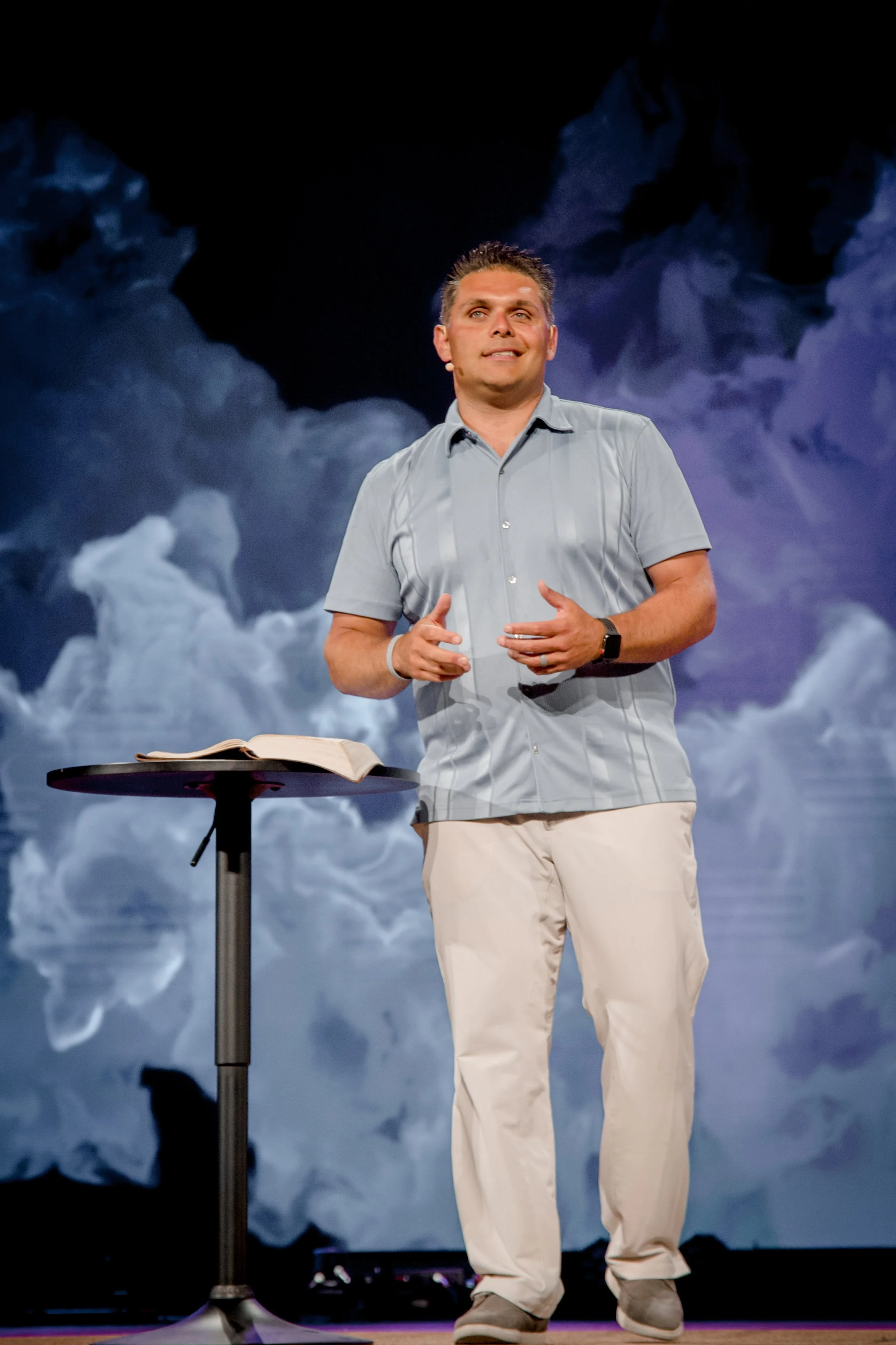 A man wearing a light gray short-sleeve shirt and khaki pants standing at a small round table with an open Bible, speaking with a backdrop of clouds.