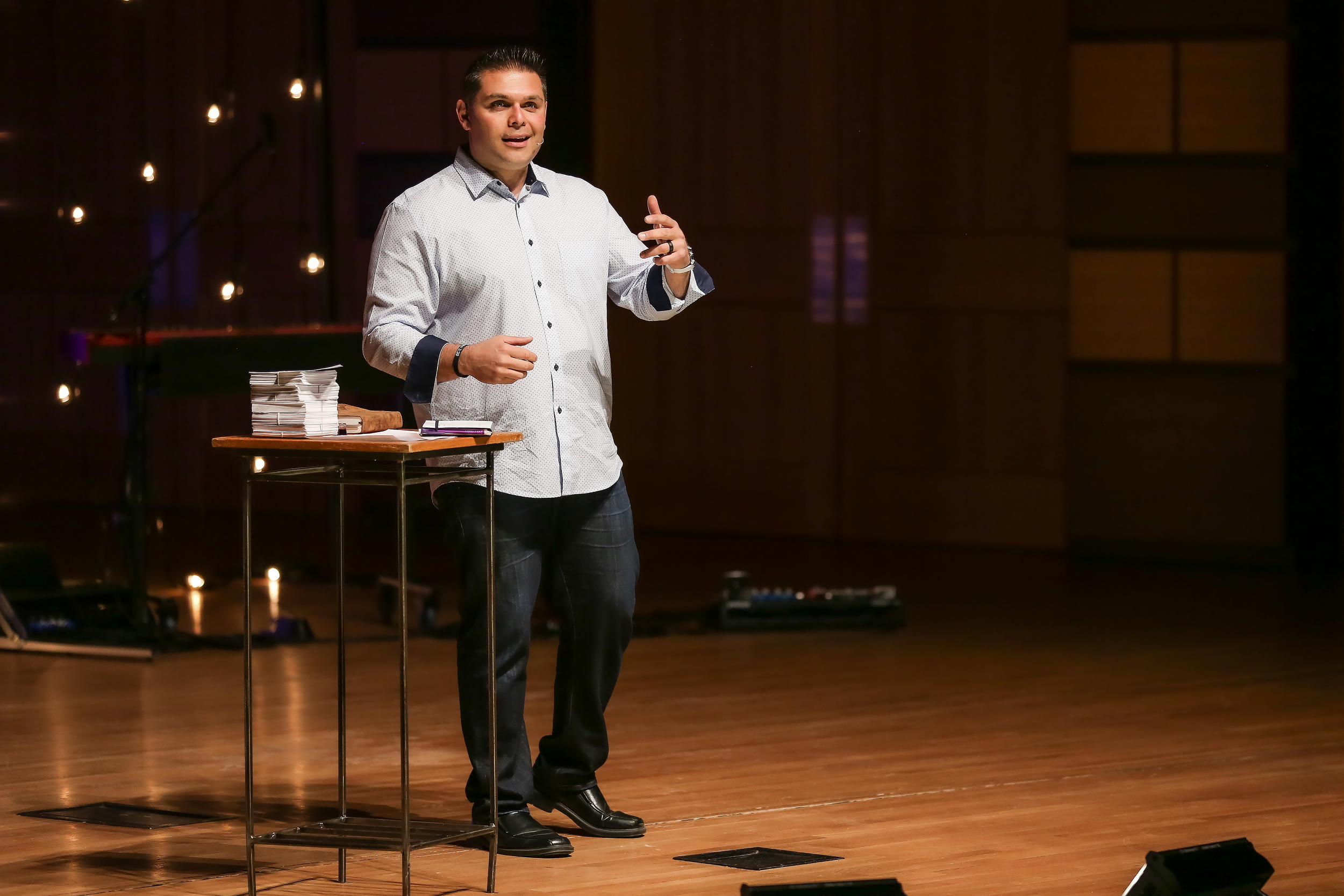 A man standing on a stage, speaking into a microphone, next to a small table with stacks of paper and a book.