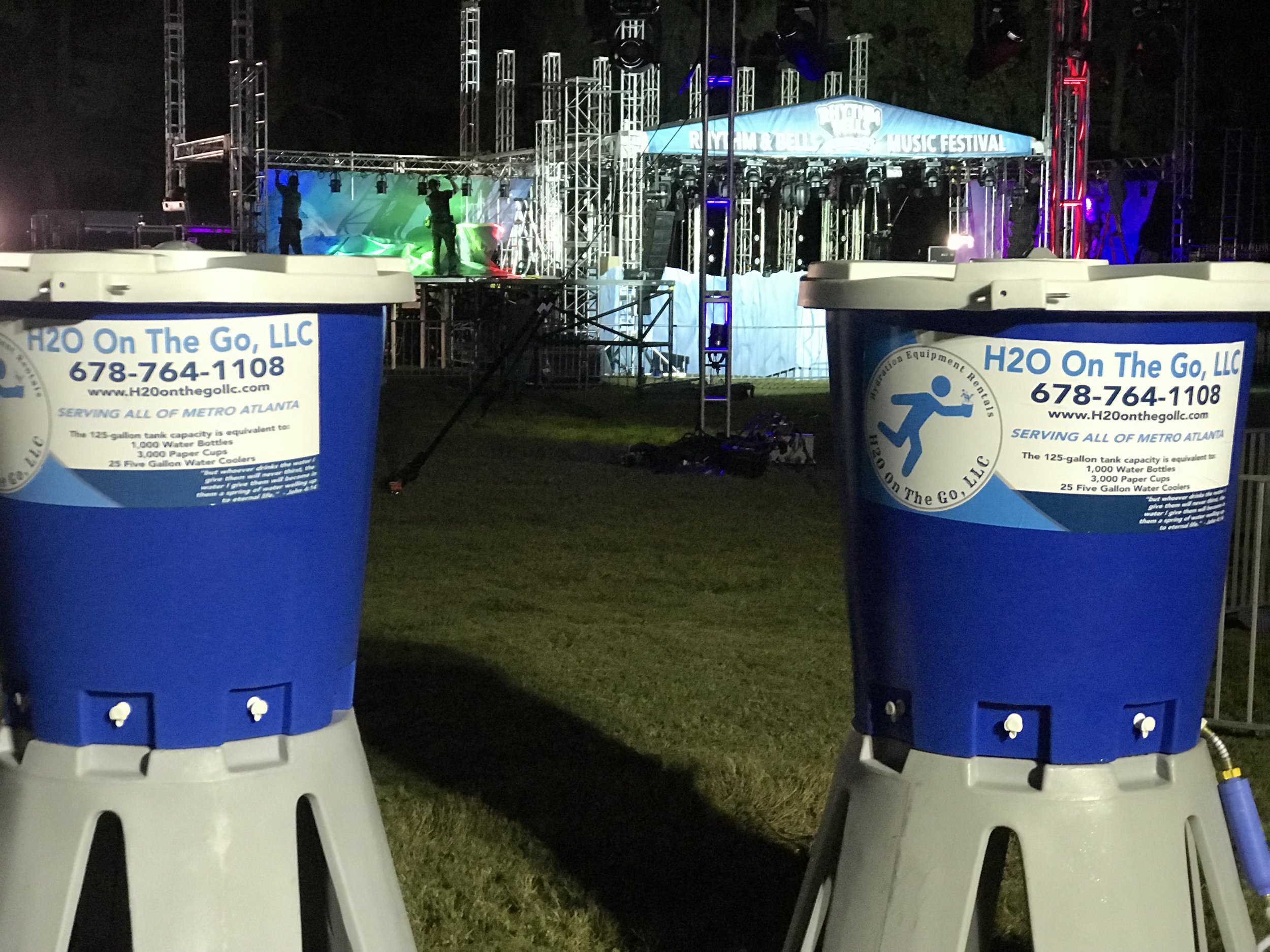 Two large blue water coolers in front of a nighttime outdoor music festival stage with lighting and sound equipment.