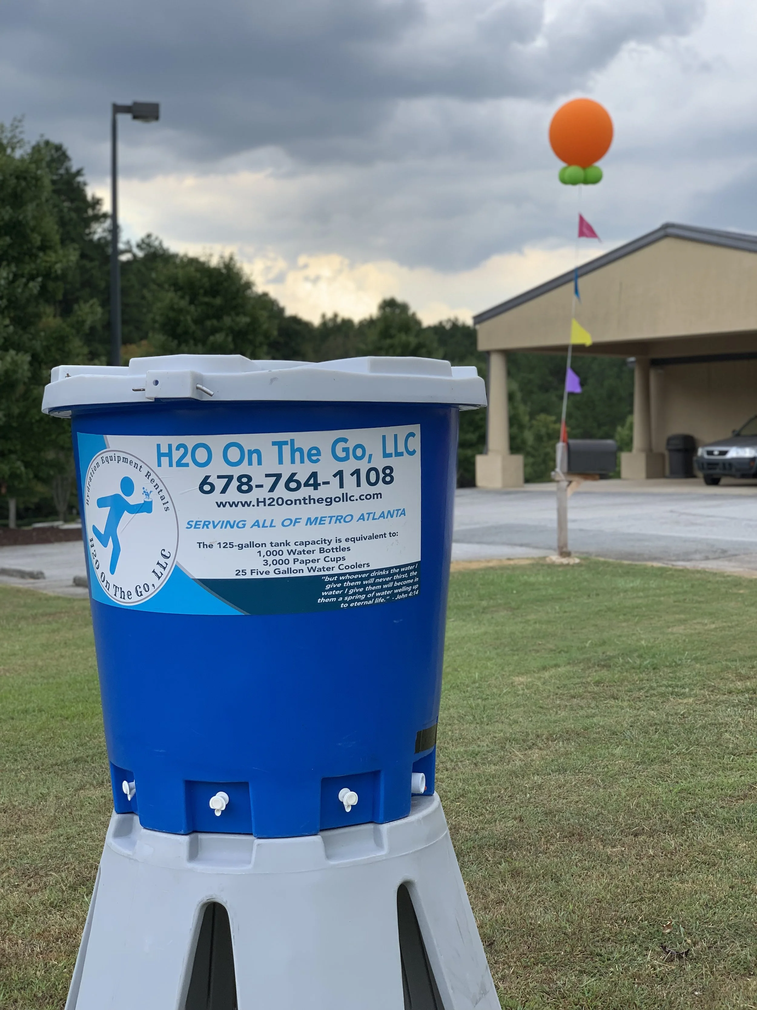 A blue water cooler with a label from H2O On The Go, LLC, placed on a grassy area outside. In the background, there's a building with a beige facade, a parking lot with a black car, a flagpole with colorful flags, a balloon decoration with orange and