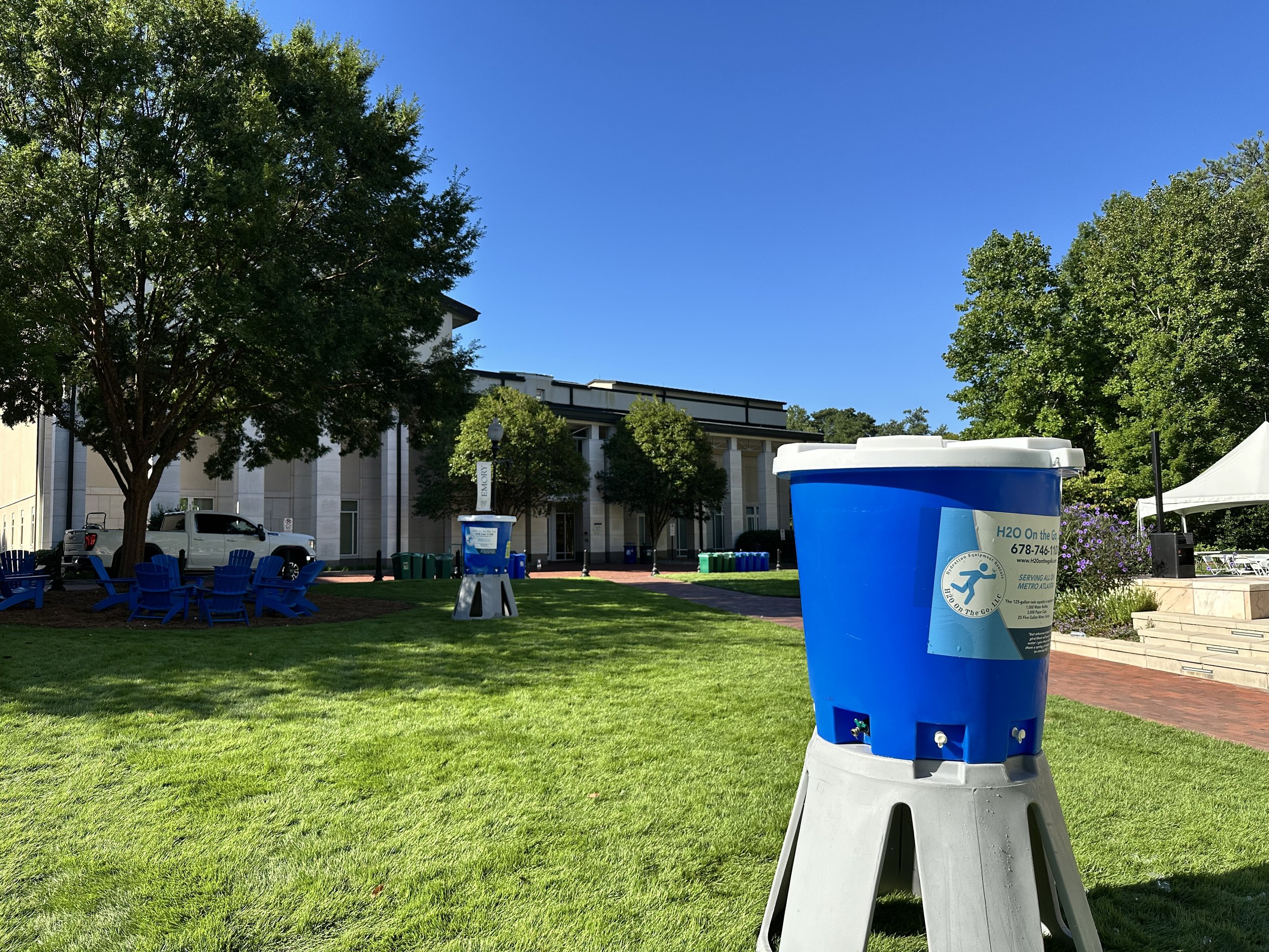 Water bottle recycling station on a lawn near trees and a building, with blue chairs and a tent in the background.