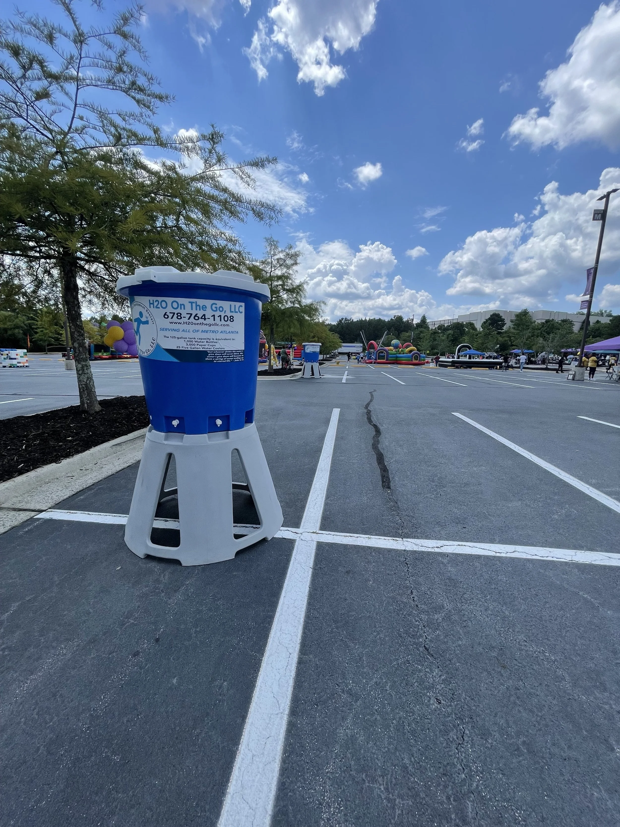 Empty outdoor parking lot with a blue water refill station, trees, and a colorful inflatable attraction in the distance under a partly cloudy sky.
