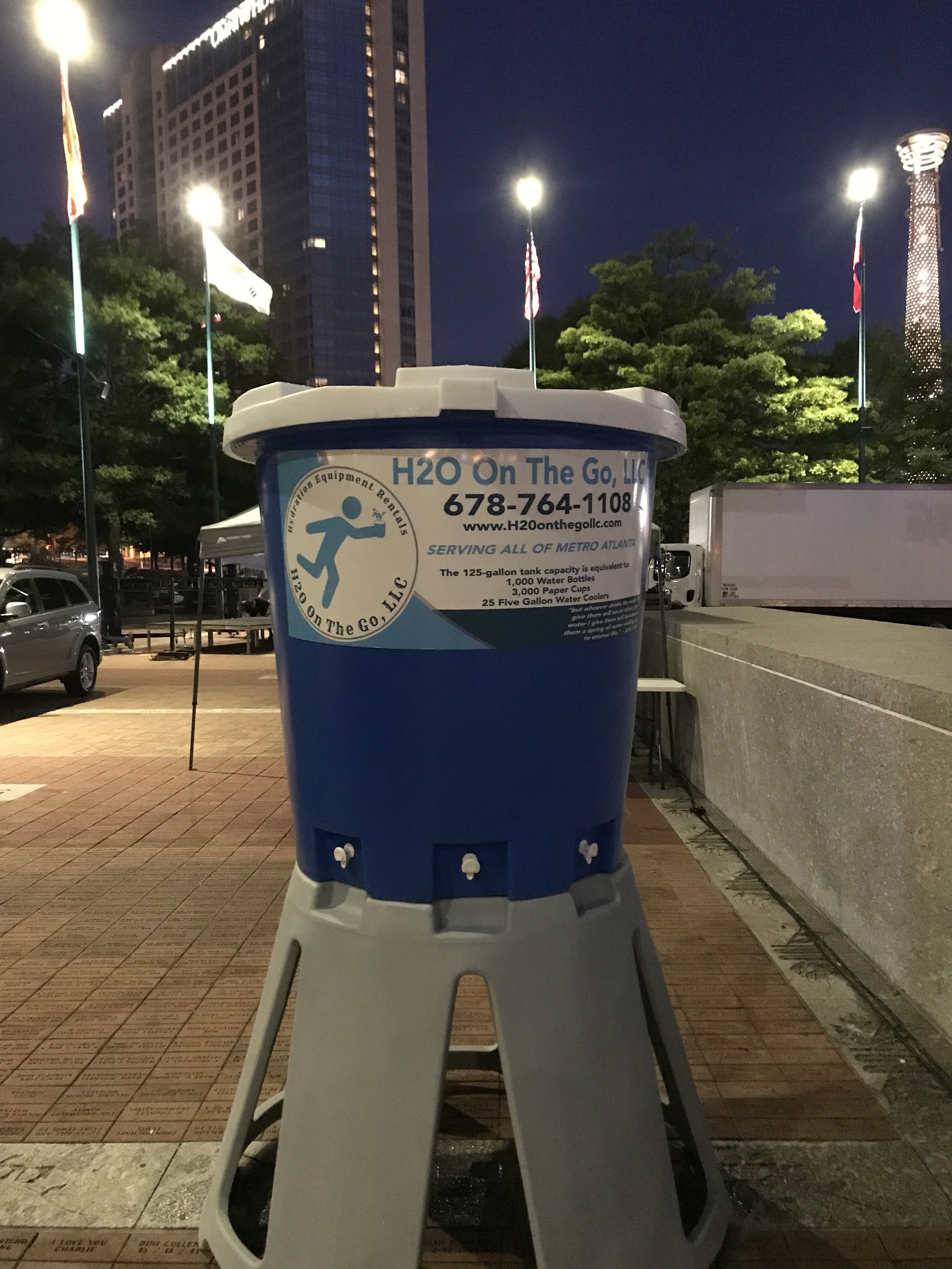 Large blue water cooler outside at night with a city skyline and flags in the background.