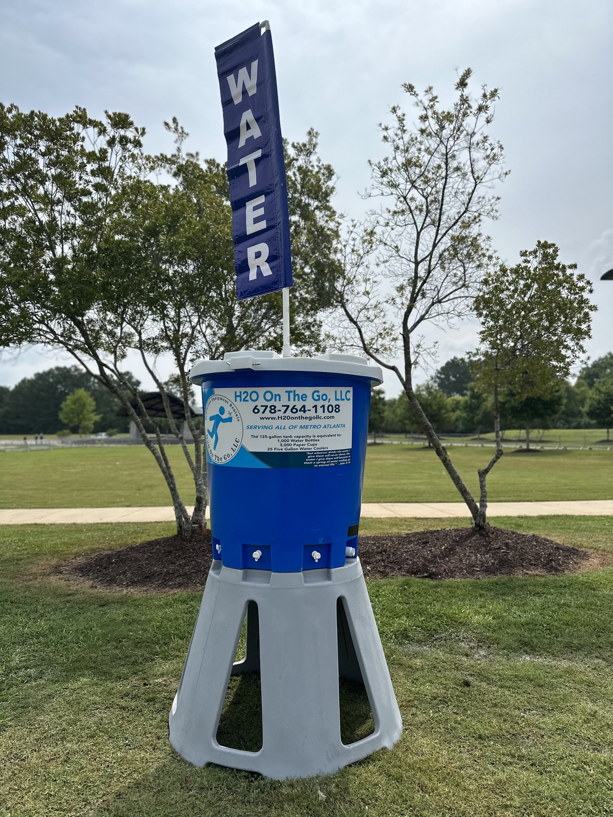 A water cooler with a blue label on a sidewalk in a park, with a flag above it that reads 'WATER' in white letters on a blue background. There are trees and grass in the background.