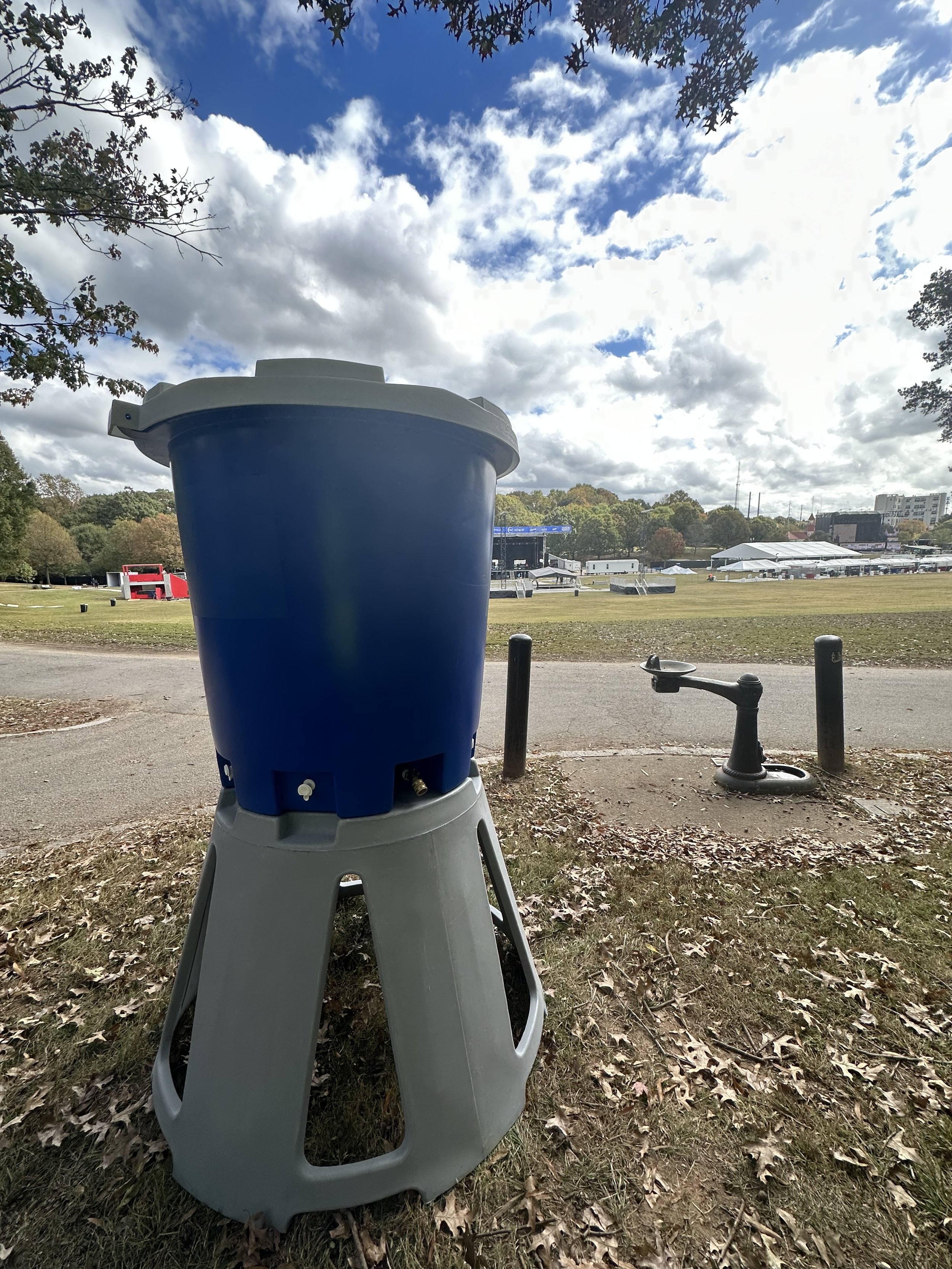 A large blue trash can on a gray stand outdoors next to a drinking fountain, with a cloudy sky and trees in the background.