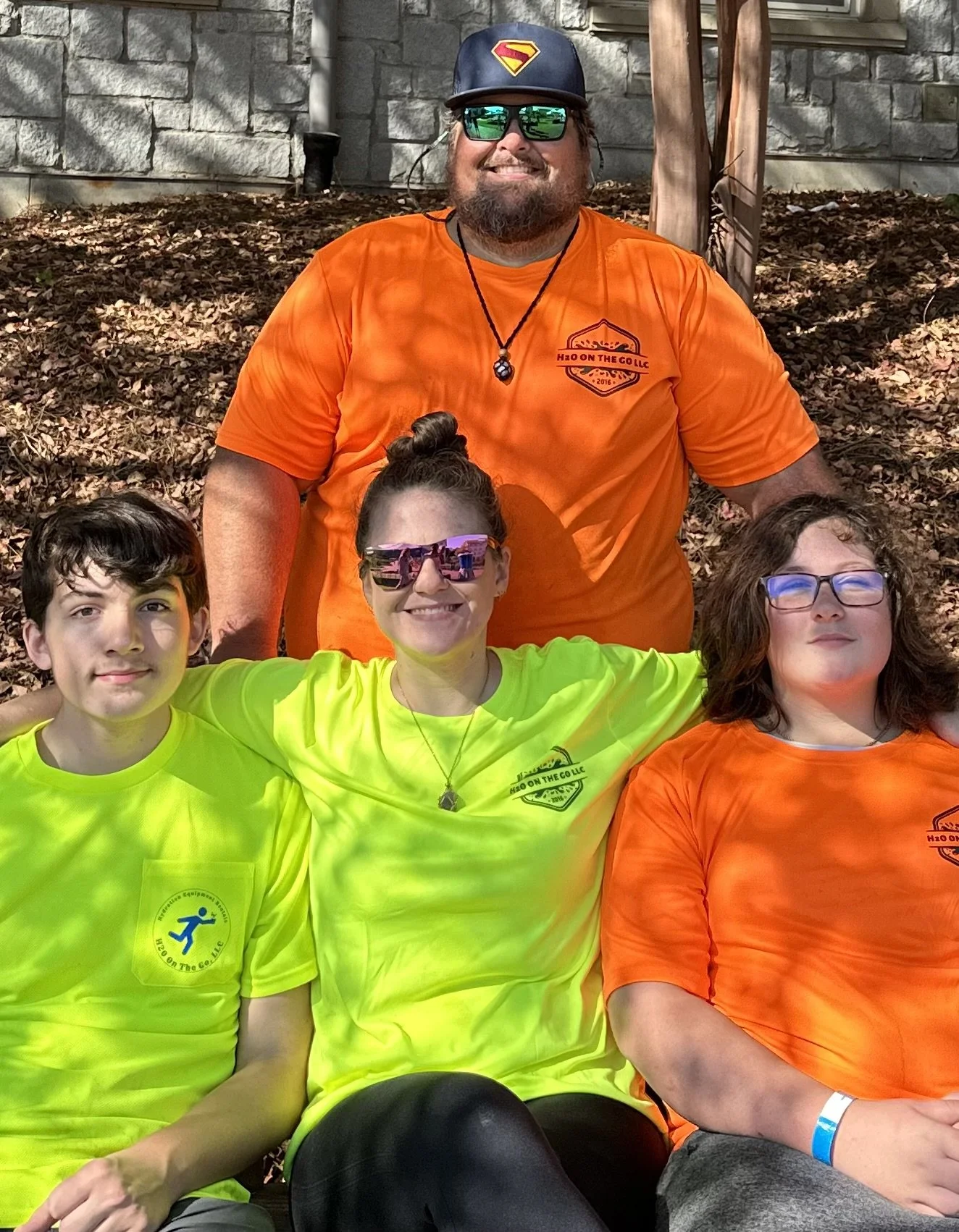 Four people sitting outdoors on a bed of leaves, with a stone wall and a wooden post in the background. The man in the back is wearing sunglasses, a cap with a Superman logo, and an orange t-shirt. The three individuals in front are wearing bright shirts, two in neon yellow and one in orange, all with matching logos.
