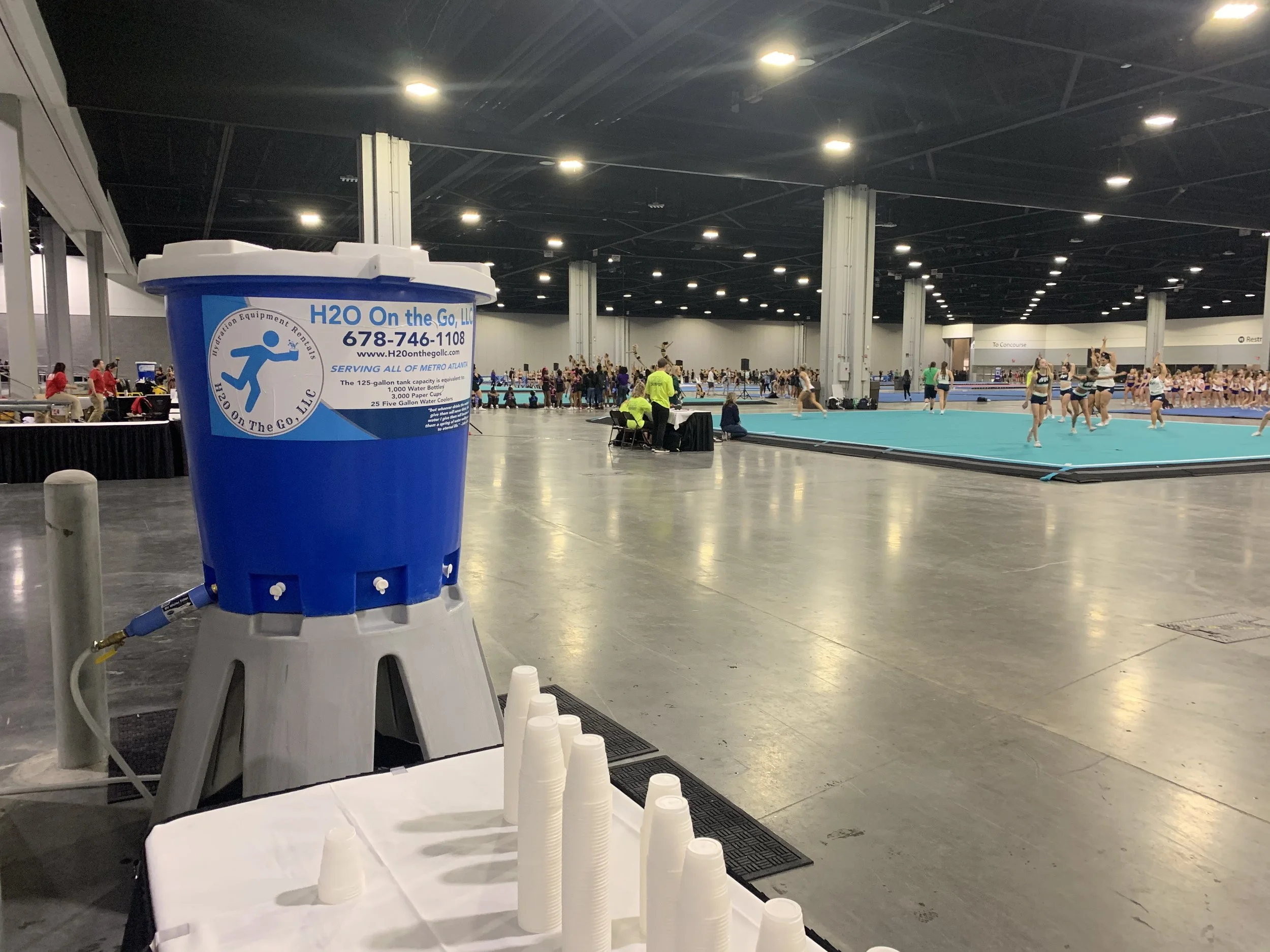 Indoor gymnastics arena with multiple groups of gymnasts practicing, a person sitting near the edge, and a water station with disposable cups in the foreground.