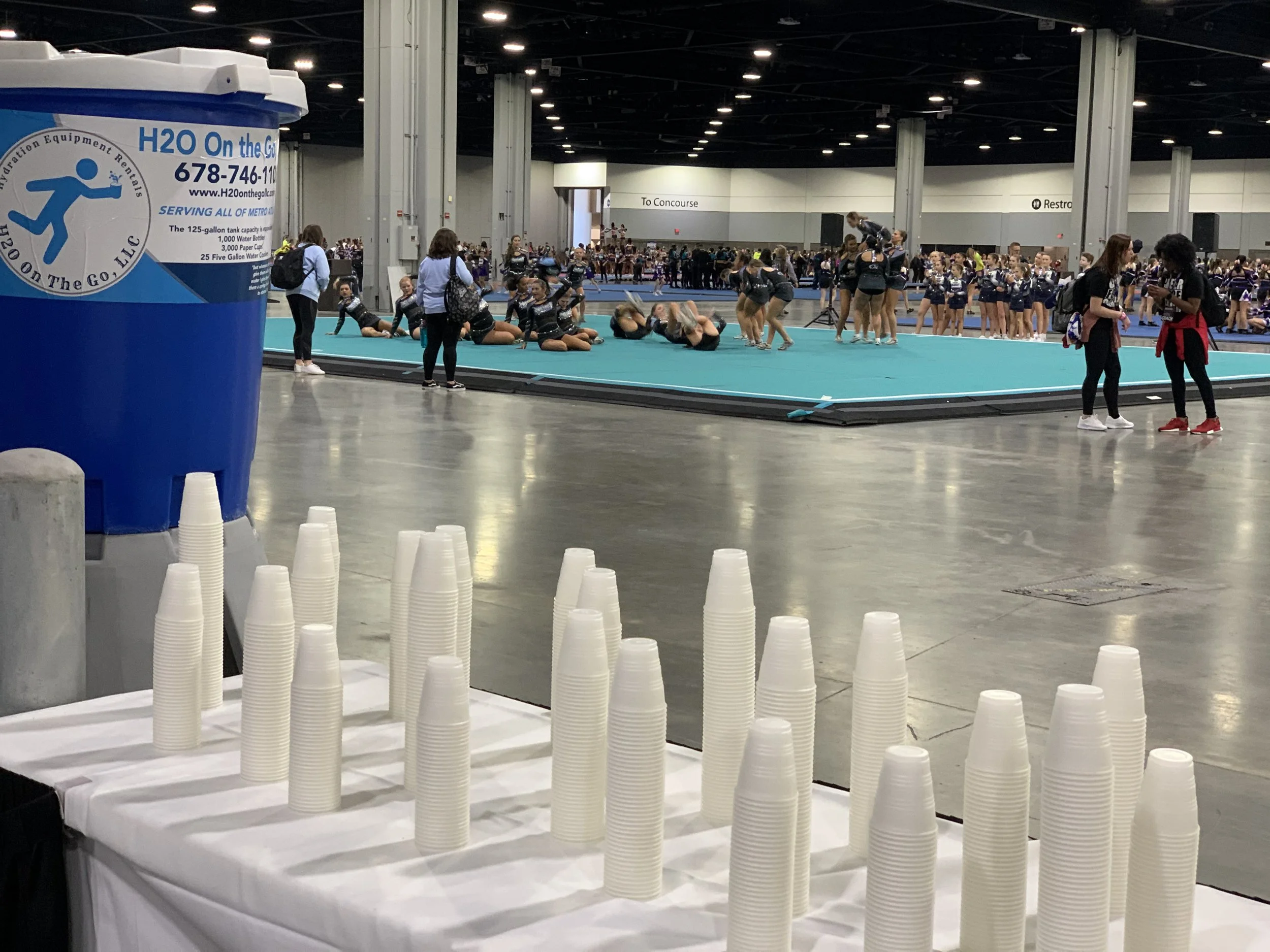 Indoor cheerleading competition with teams practicing on mats, spectators and staff observing, a water station with stacks of plastic cups in the foreground, and a large open space with high ceilings.