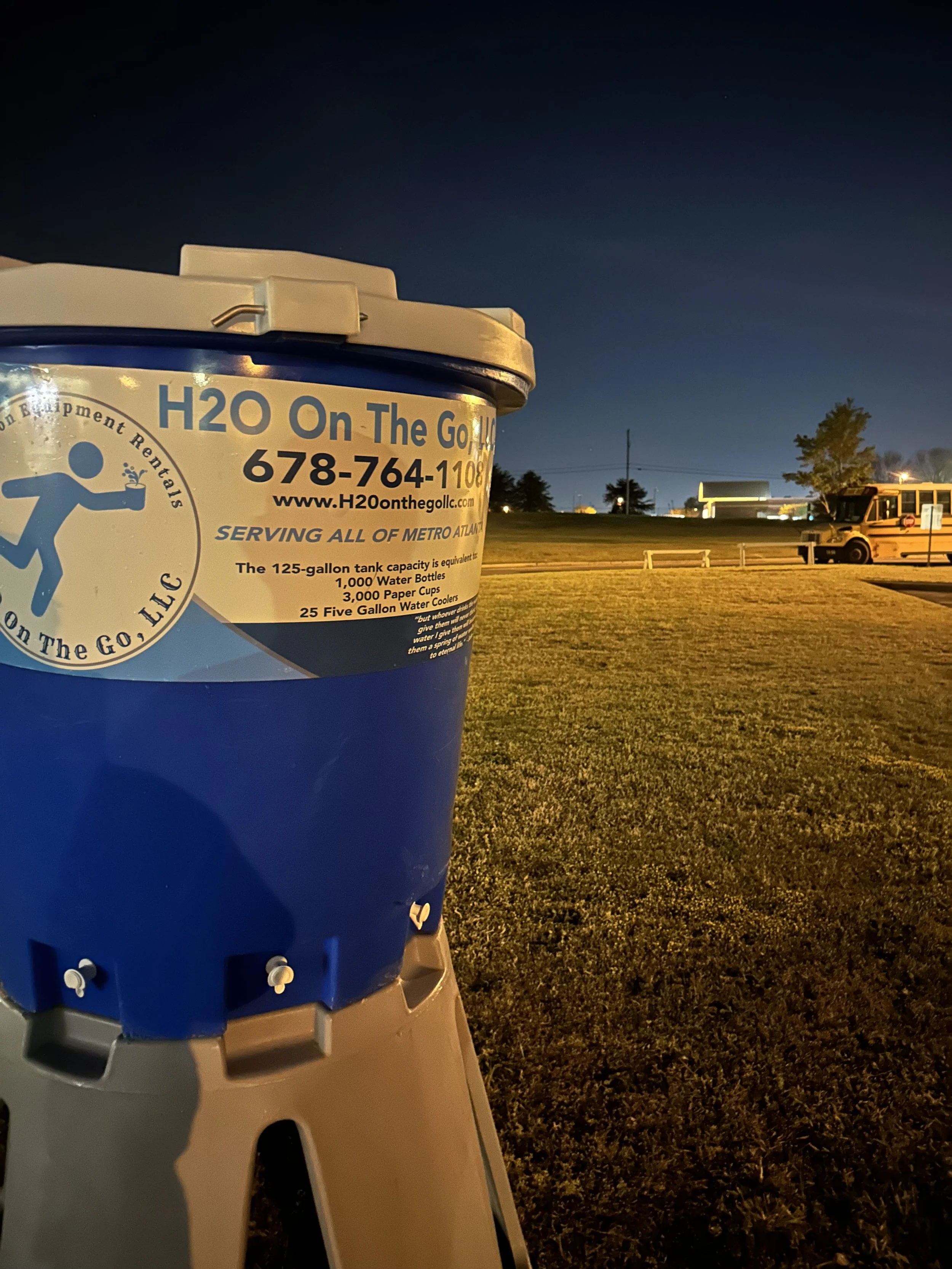 A blue water cooler jug with a label advertising water rental services, placed outdoors at night with a grassy field and a yellow school bus in the background.